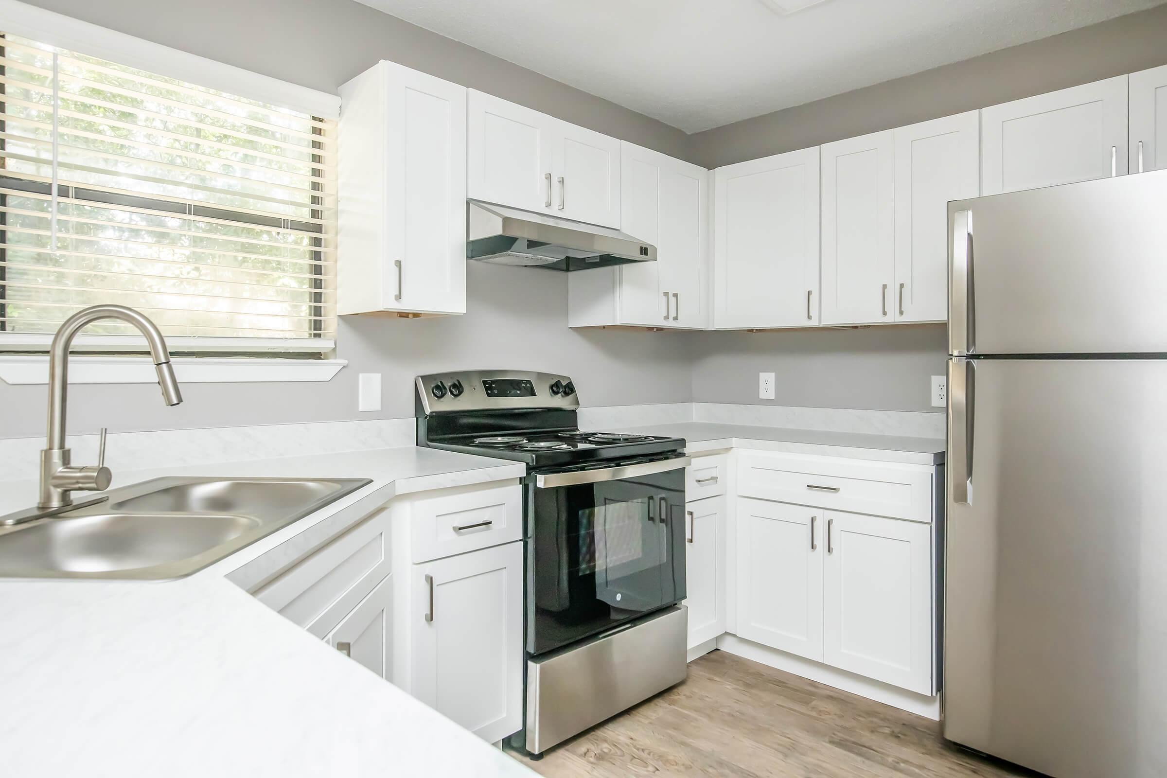 Modern kitchen featuring white cabinets, a stainless steel refrigerator, a black stove, and a double sink. Natural light streams through a window with blinds, highlighting the clean lines and contemporary design of the space. The flooring is a light wood-tone, enhancing the bright and airy atmosphere.