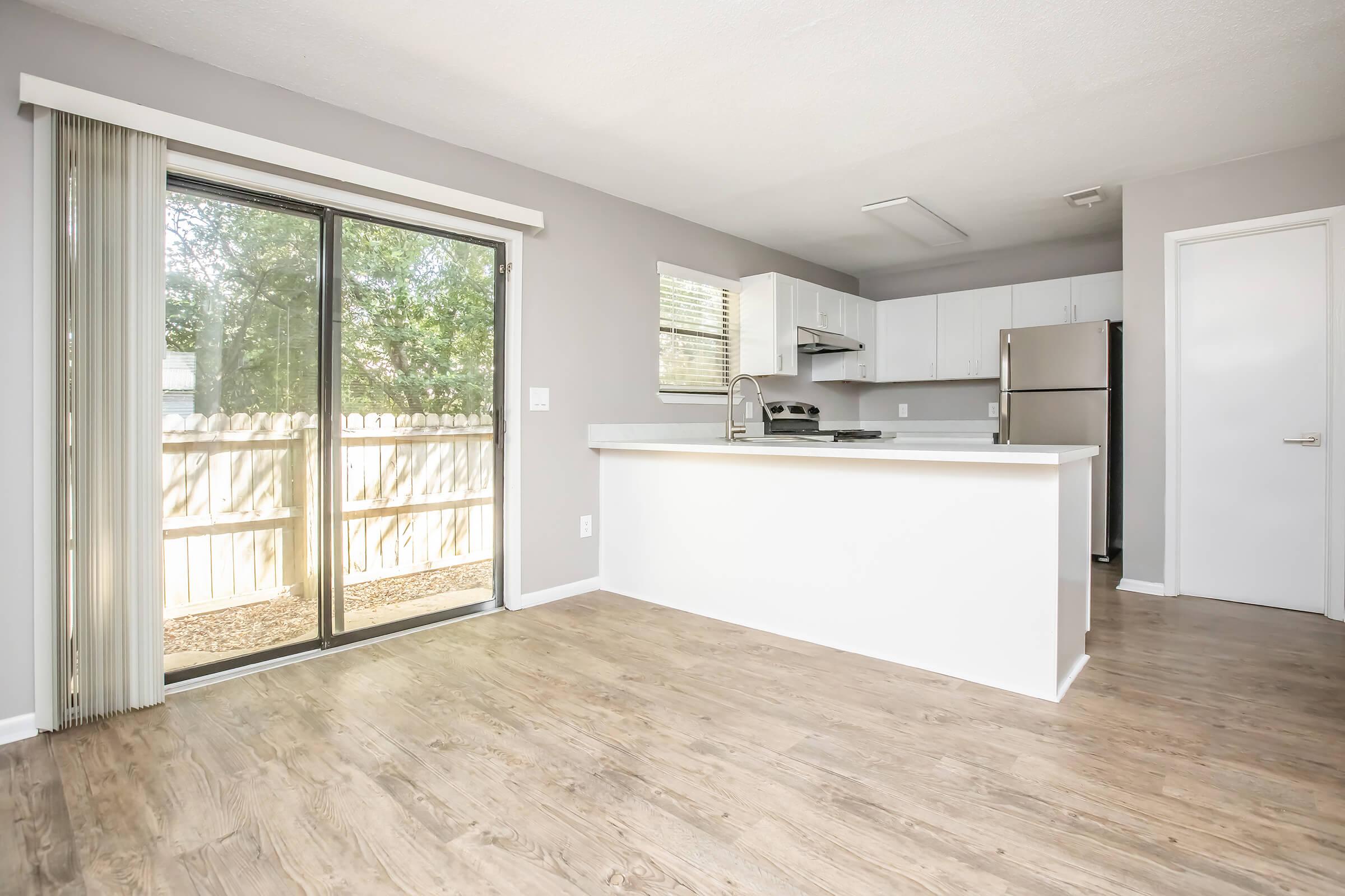 A modern kitchen and living area featuring a sliding glass door that opens to a yard, light-colored walls, a kitchen island with a sink, and stainless steel appliances. Large windows allow natural light to brighten the space, with wooden flooring throughout.