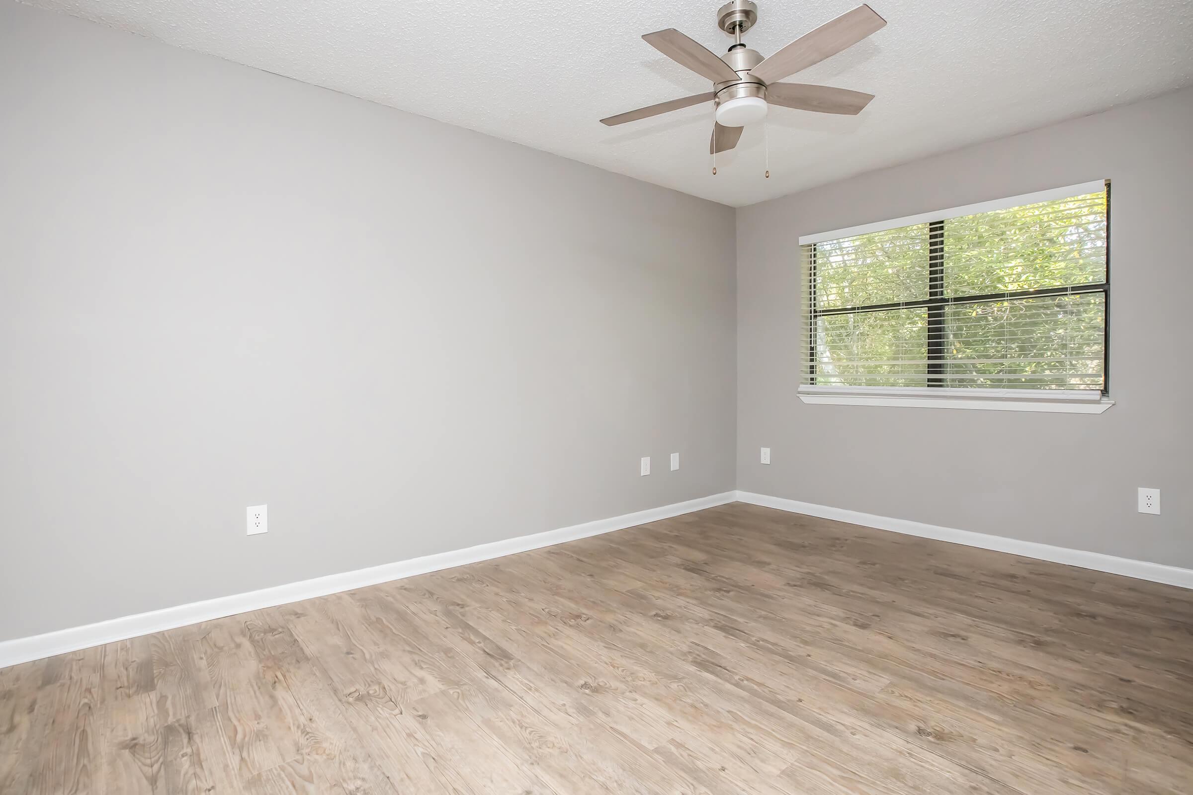 A simple, empty room with light gray walls and a ceiling fan. The floor features a light wood finish. A window with blinds allows natural light to enter, providing a view of greenery outside. The room has no furniture, creating a spacious feel.