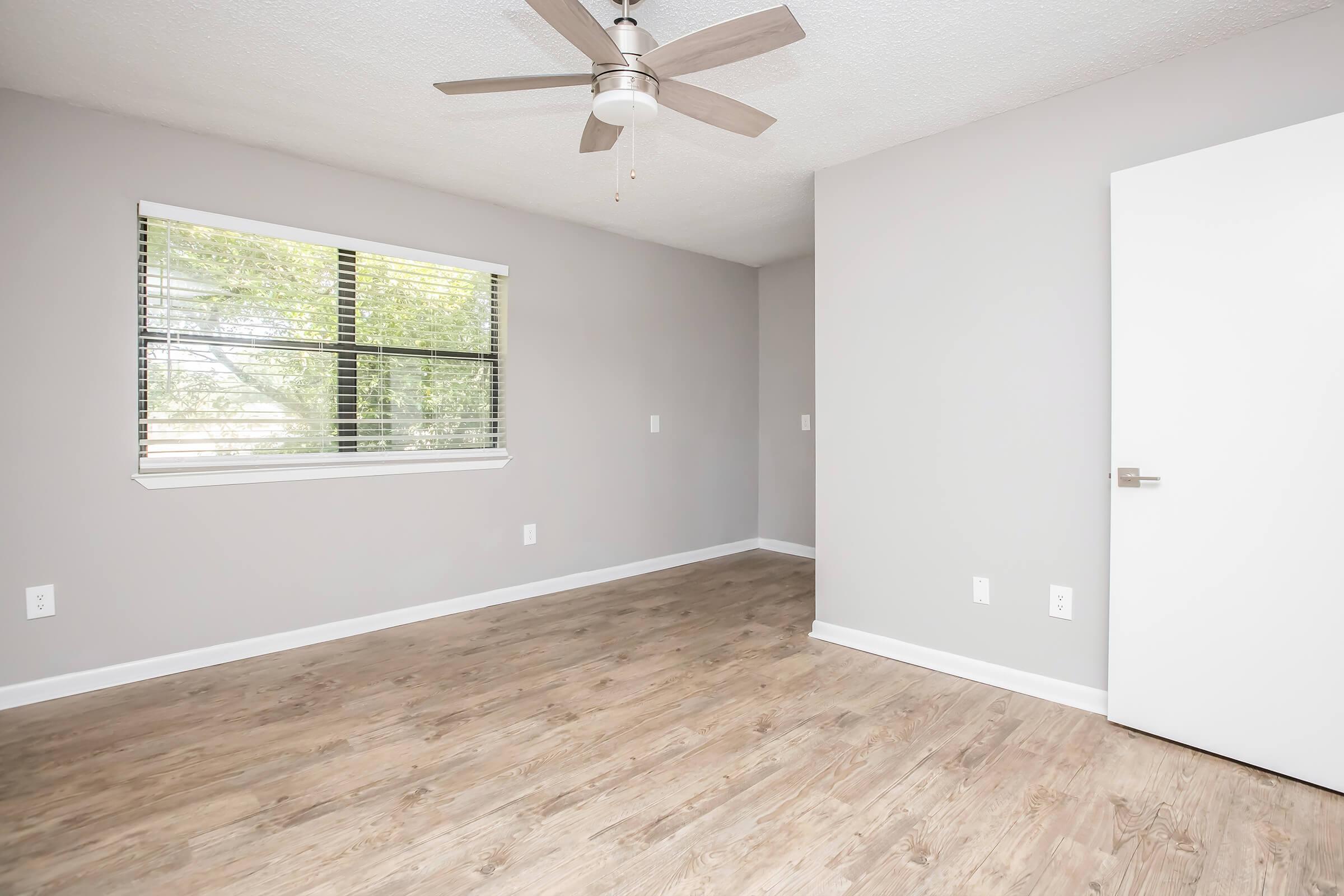 A spacious, empty room with light gray walls and a ceiling fan. It features a window with blinds, allowing natural light to enter. The floor is covered in light-colored wood laminate, and there’s a modern white door on the right side. The overall atmosphere is bright and minimalistic.