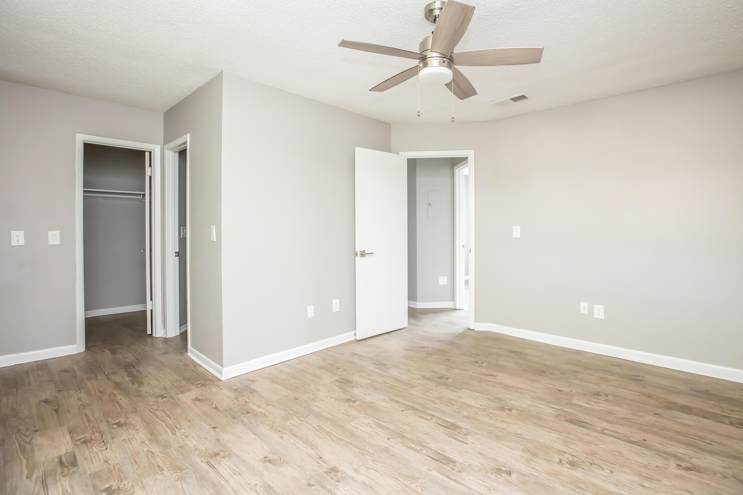 A light and airy empty bedroom with gray walls and wood-like flooring. A ceiling fan is mounted on the ceiling. There are two doorways leading to adjoining rooms, one of which features a closet. The space is well-lit and appears inviting, ideal for furnishing and personal decoration.
