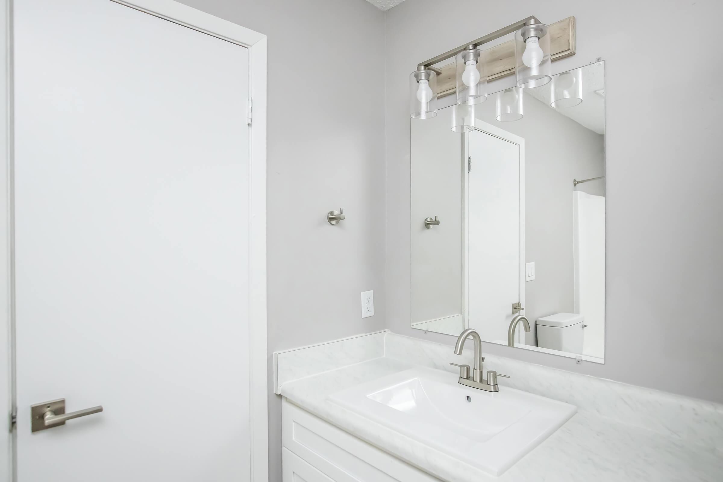 A modern bathroom featuring a white countertop with a sink, a large mirror above, and contemporary light fixtures. The walls are painted in a light gray color, and there is a door leading to another room. A toilet is visible in the background. The overall design is clean and minimalist.