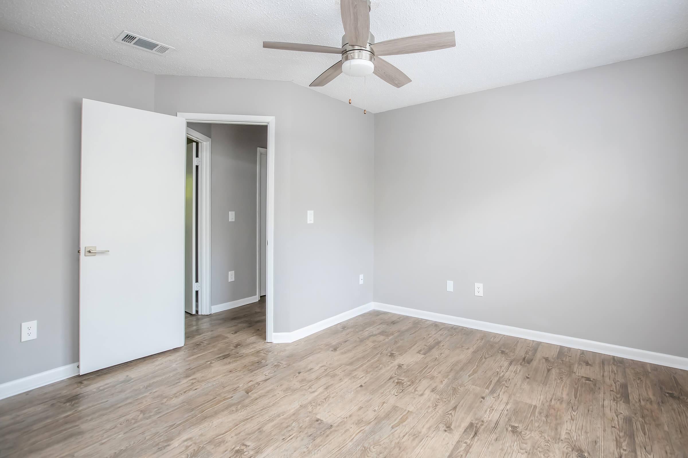 A well-lit, empty room featuring a light gray wall and wooden flooring. A ceiling fan is mounted on the ceiling, and there is an open white door leading to another room on the left. The space is clean and modern, creating a fresh and inviting atmosphere.