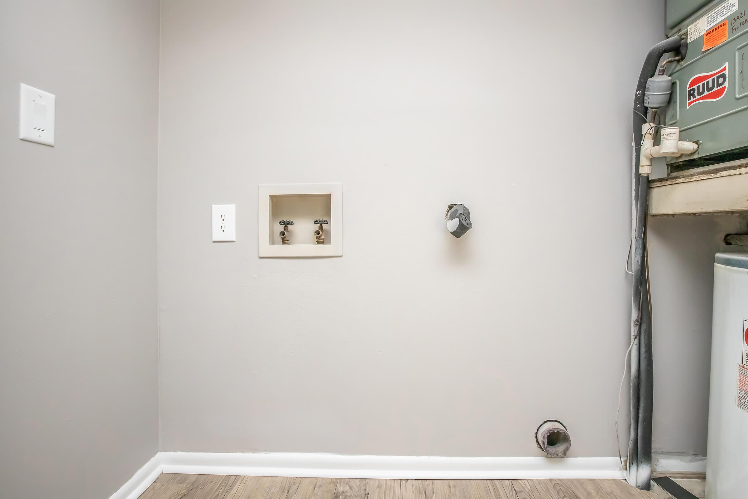 Interior wall with a light switch and a water supply box featuring two taps. A single faucet connection is visible, along with a grey pipe running from the side, and a water heater partially seen in the corner. The floor is covered with light-colored laminate.