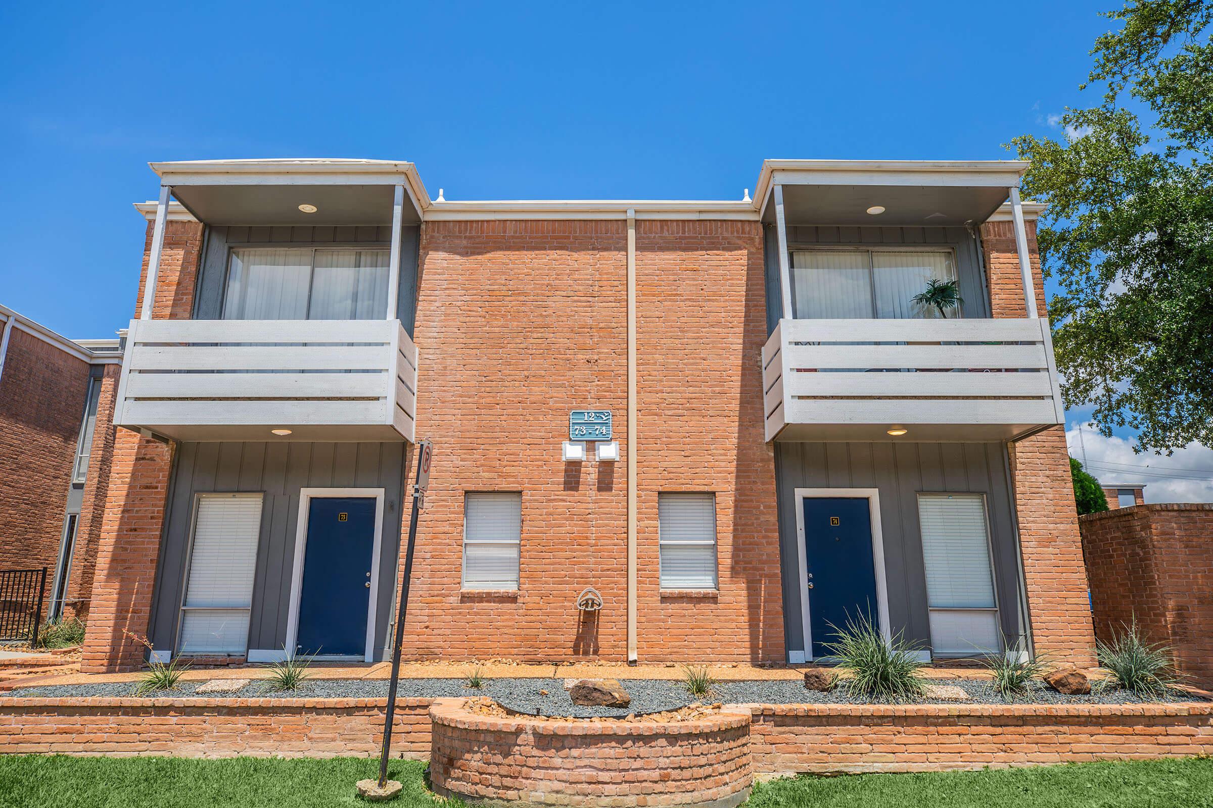 Two-story brick apartment building with a modern design. Each unit features a small balcony and front door, framed by decorative landscaping with plants and rocks. The blue sky and sunlight highlight the building's architecture.