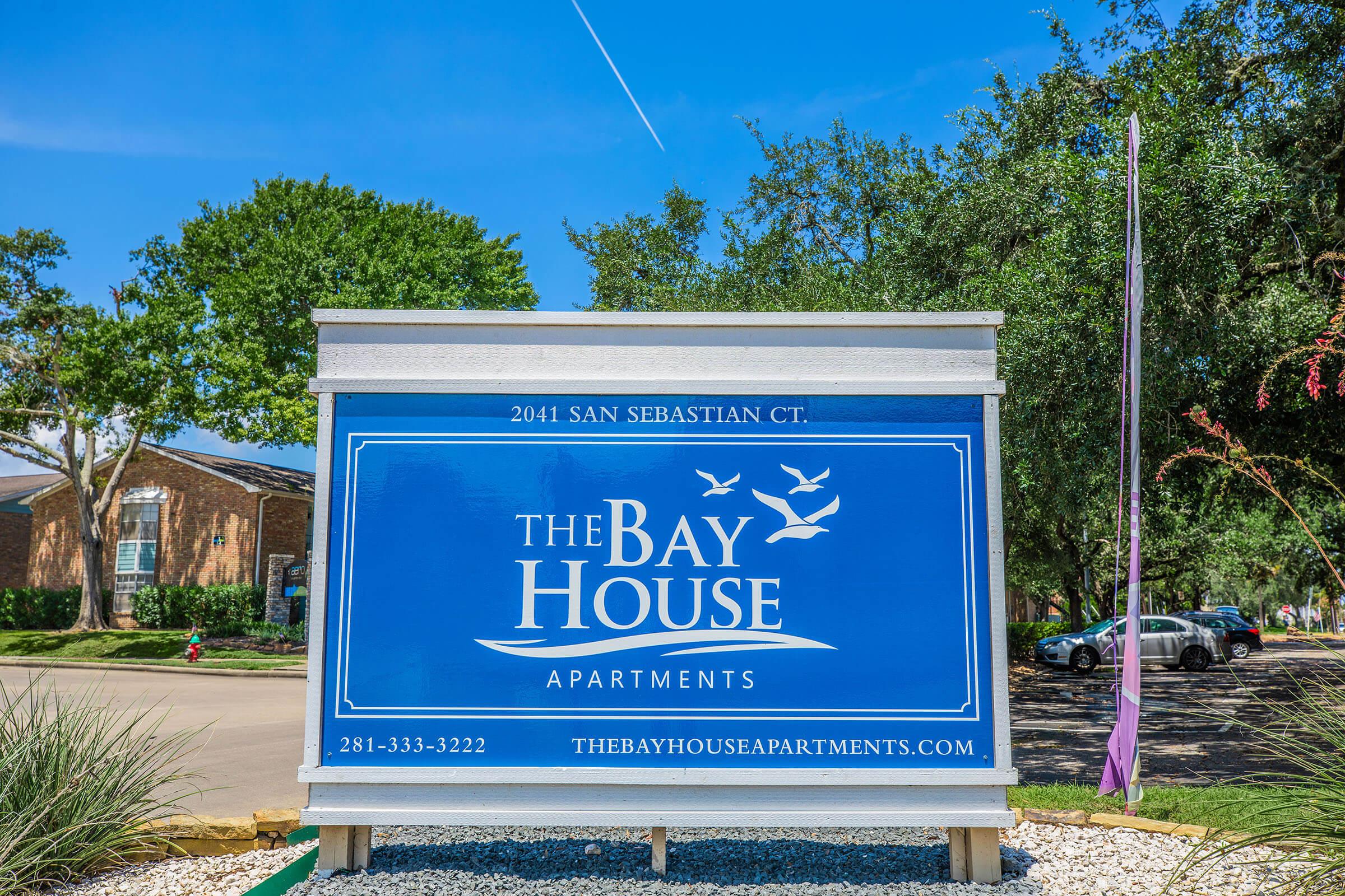 A large, blue sign for The Bay House Apartments, displaying the address "2041 San Sebastian Ct." and the phone number "281-333-3222." The sign features a logo with birds and is set against a clear blue sky with some trees in the background.