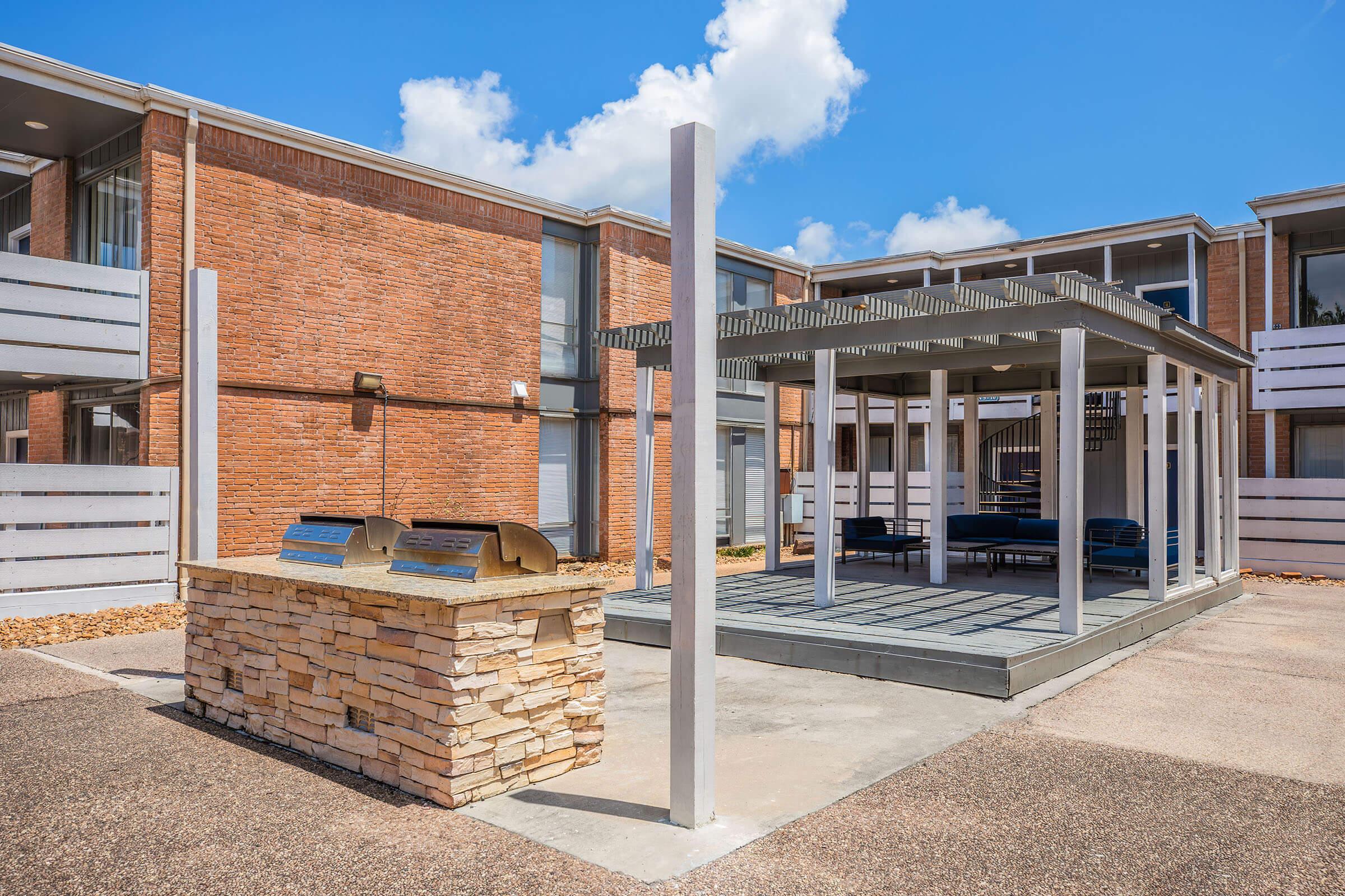 Outdoor patio area featuring a stone grill station, wooden pergola, and seating beneath an open sky. The surrounding buildings are made of red brick and have modern architectural elements, with a clean and inviting atmosphere.