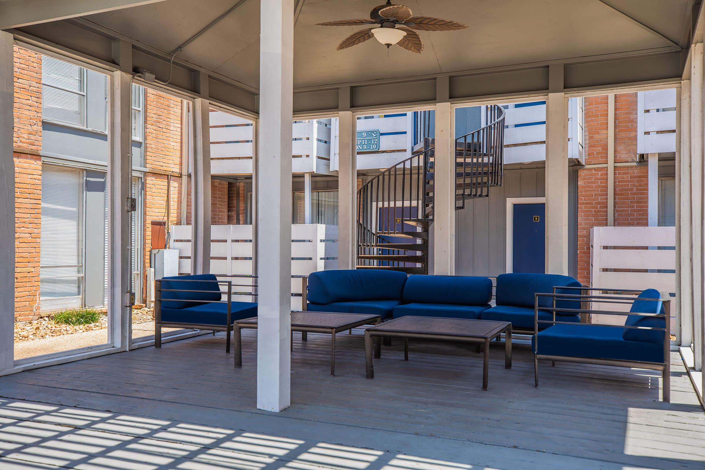 A spacious covered patio area featuring a blue sectional sofa and a coffee table. The setting includes large windows and a view of a spiral staircase nearby, with brick building exteriors in the background.