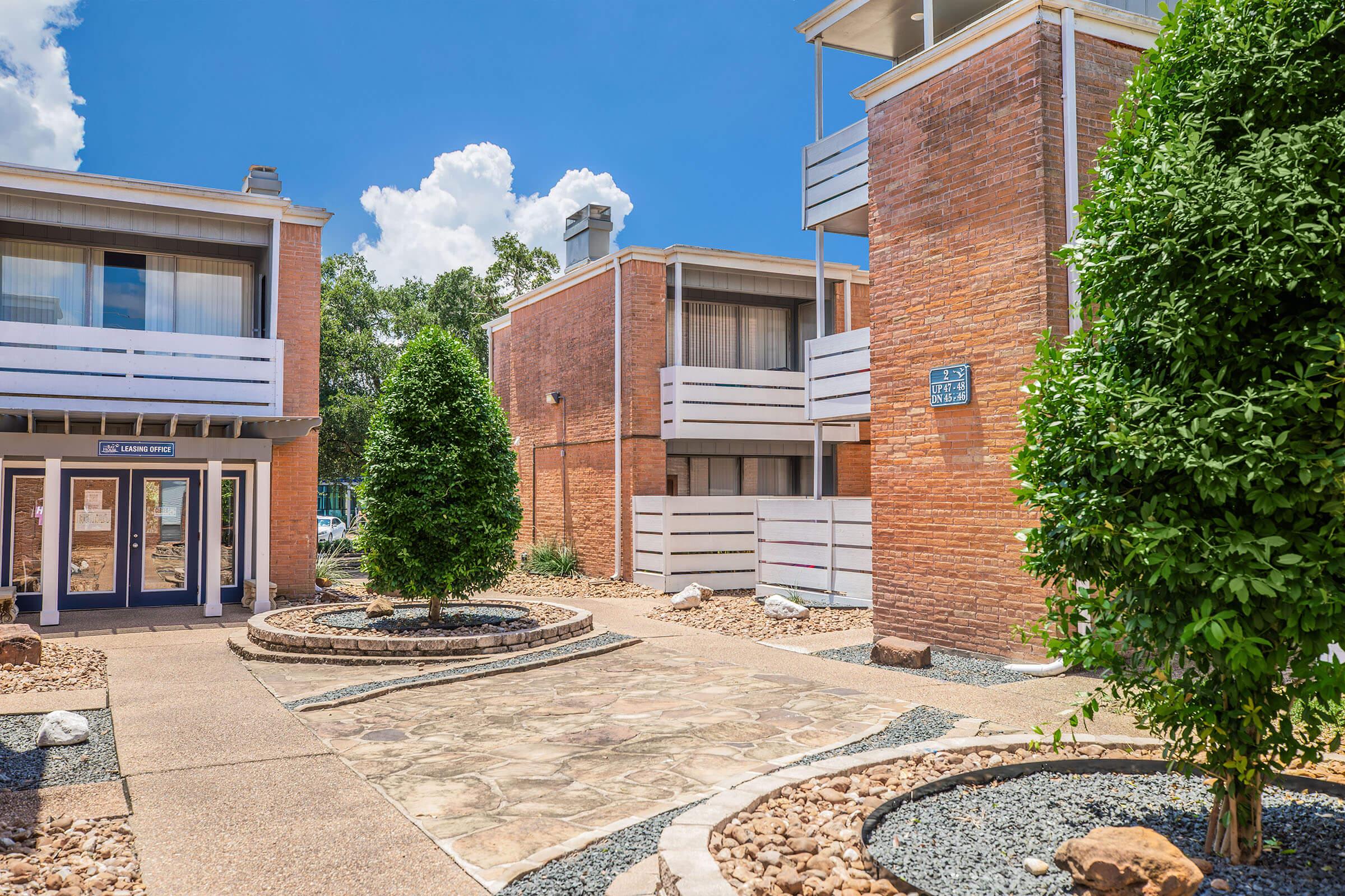 A view of a landscaped area in front of two residential buildings. The pathway features a stone surface bordered by small rocks and greenery. The buildings are constructed of brick, with balconies visible. The sky is bright and blue with a few clouds.