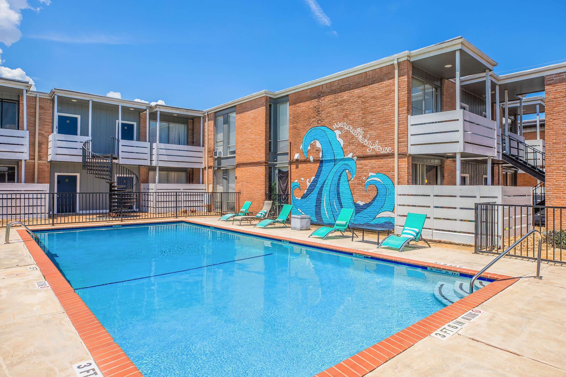 A bright blue swimming pool surrounded by lounge chairs, with a vibrant wave mural on the brick wall nearby. The pool area is framed by two-story apartment buildings under a clear blue sky, creating a welcoming and refreshing outdoor atmosphere.