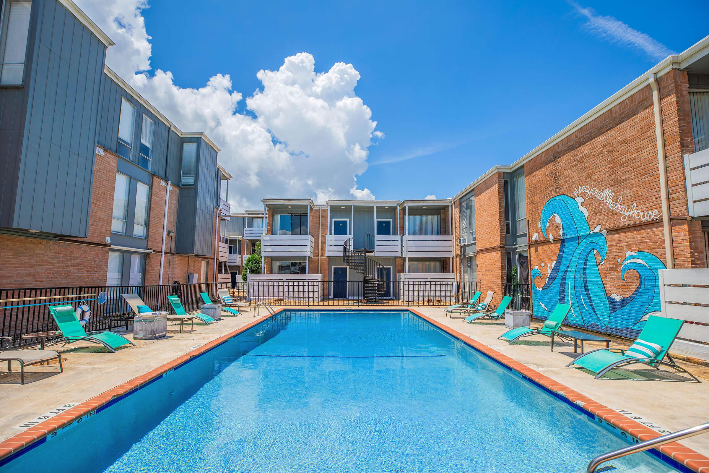 A bright and inviting swimming pool surrounded by lounge chairs, located in a courtyard of multi-story apartment buildings. The sky is partly cloudy with vibrant blue tones, and a colorful wave mural decorates one of the buildings nearby. The scene is relaxing and sunny, ideal for leisure activities.