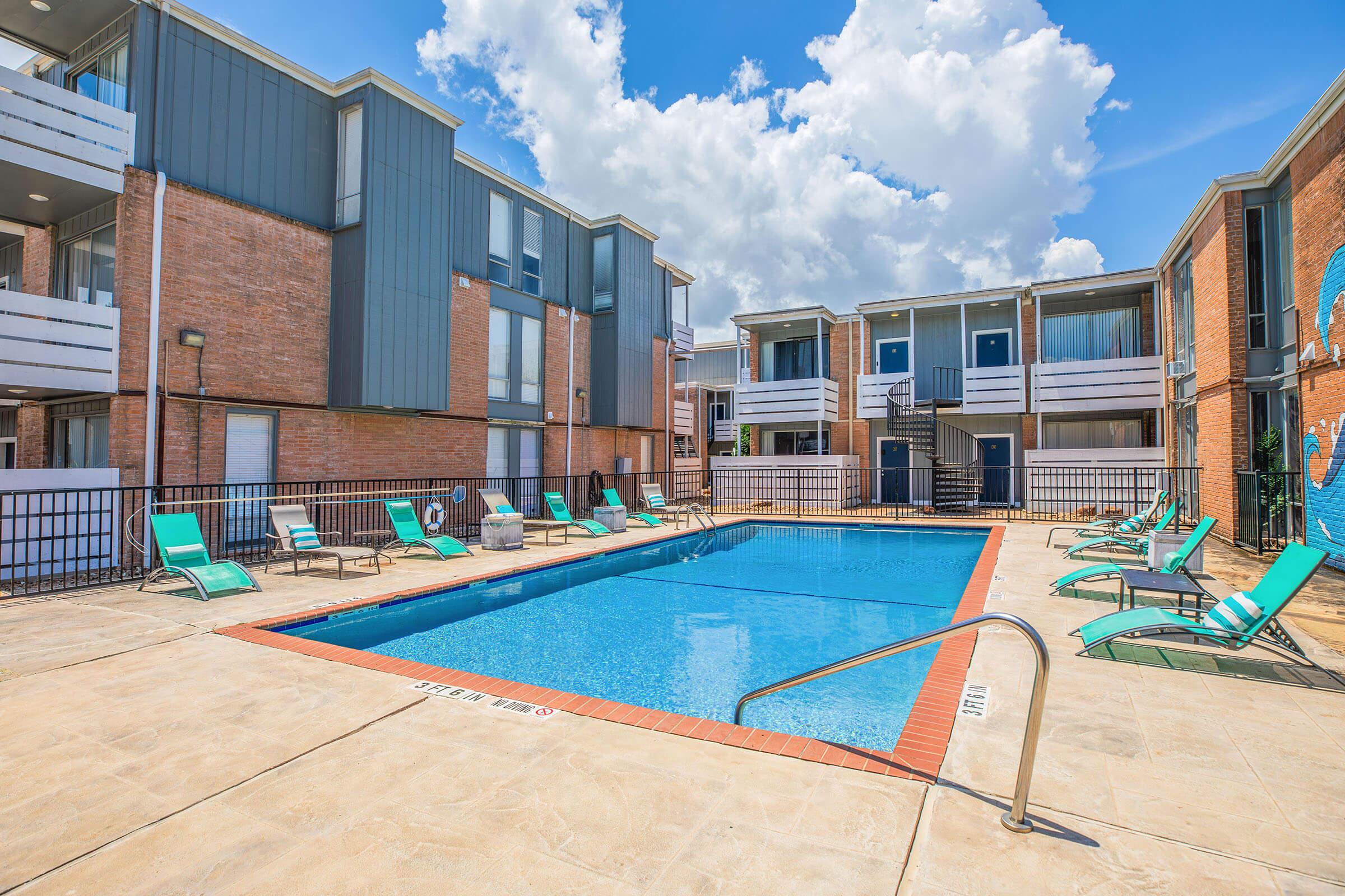 A rectangular swimming pool surrounded by lounge chairs and a fenced area, with two stories of apartment buildings in the background. The sky is partly cloudy, and the scene is well-lit, conveying a relaxed atmosphere.