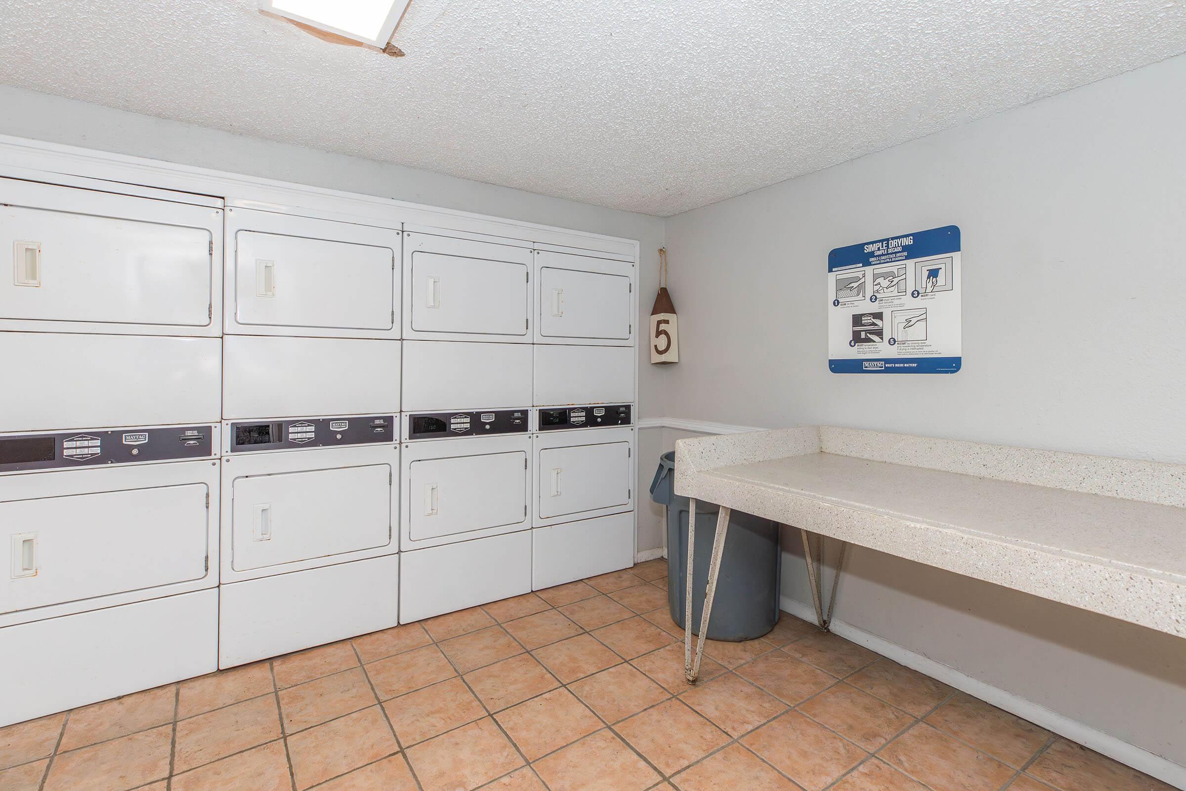 A clean laundry room featuring a row of white washing machines and dryers against a light grey wall. There is a folding table made of speckled laminate, a trash bin, and a wall-mounted sign with laundry instructions. The floor is tiled with orange-brown squares.