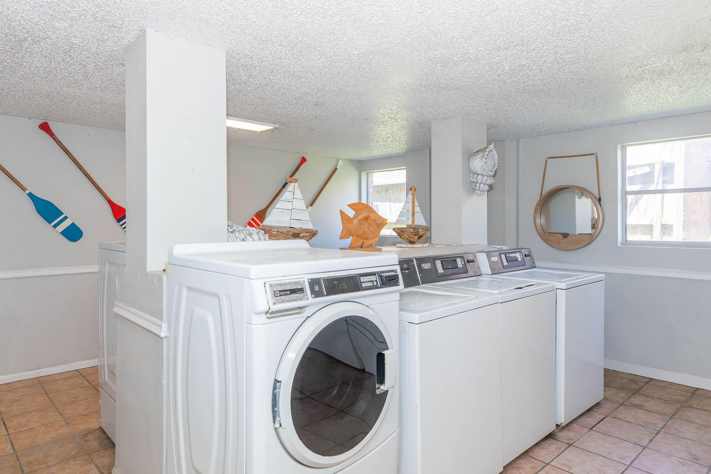 Bright laundry room featuring two washing machines and a dryer, with decorative items like wooden paddles, a sailboat model, and a fish sculpture. Natural light comes through the windows, highlighting the tiled floor and clean, white walls.