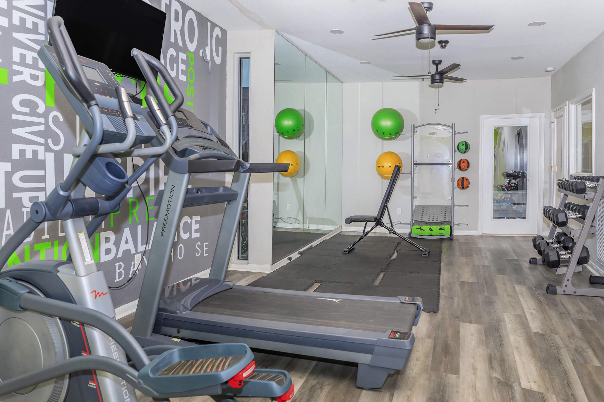 A modern home gym featuring a treadmill, weights, and exercise equipment. The walls are decorated with motivational phrases, and there are colorful exercise balls on one side. Large mirrors enhance the space, and the flooring is a mix of tiles and wood. Ceiling fans provide ventilation.