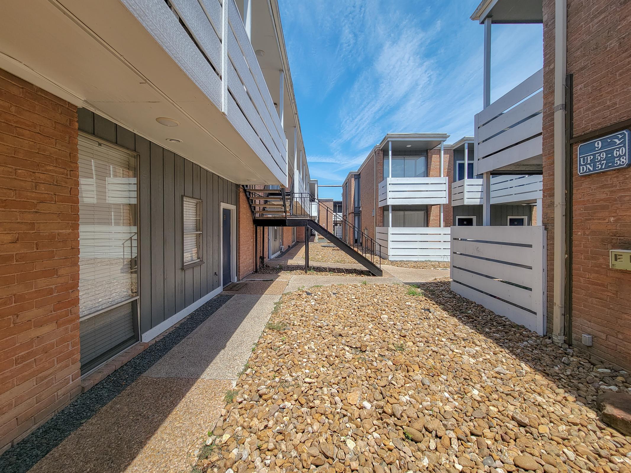 View of a residential courtyard featuring two blocks of apartments with a gravel pathway in between. An exterior staircase connects the floors of one building. The sky is blue with scattered clouds, and the buildings have a mix of brick and wooden facades.
