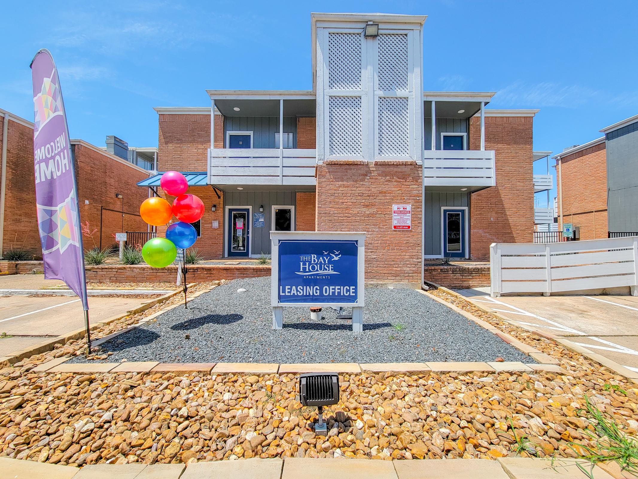 Leasing office of The Bay House, featuring a brick building with white accents. In front, a sign reads "Leasing Office," accompanied by colorful balloons and a welcoming banner. The area is landscaped with gravel and stones, under a clear blue sky.