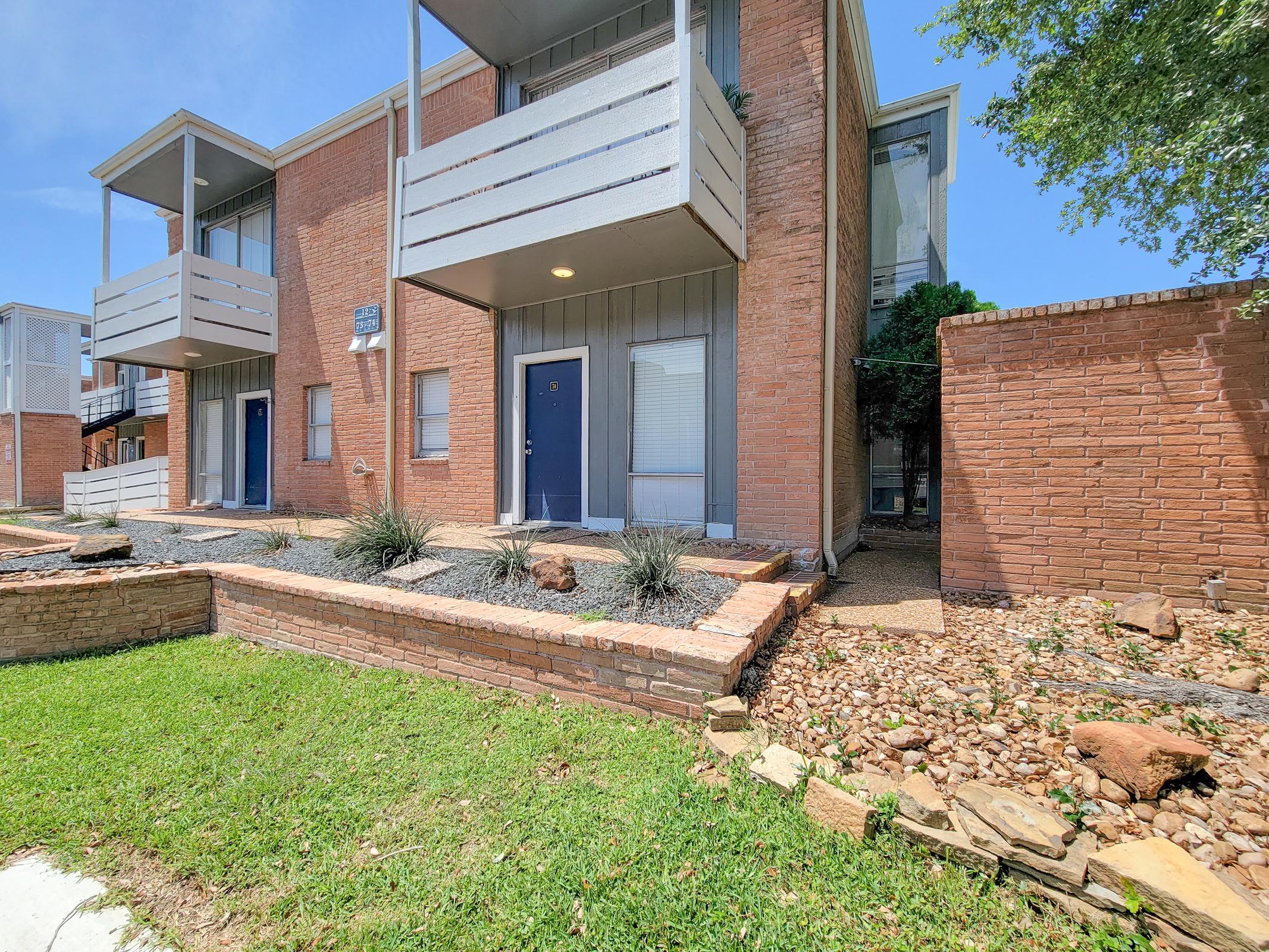 A modern brick apartment building featuring two stories, blue doors, and white balconies. The surrounding area includes a landscaped lawn with rocks and low shrubs, and a small stone pathway leading to the entrance. The sky is clear and blue, indicating a sunny day.