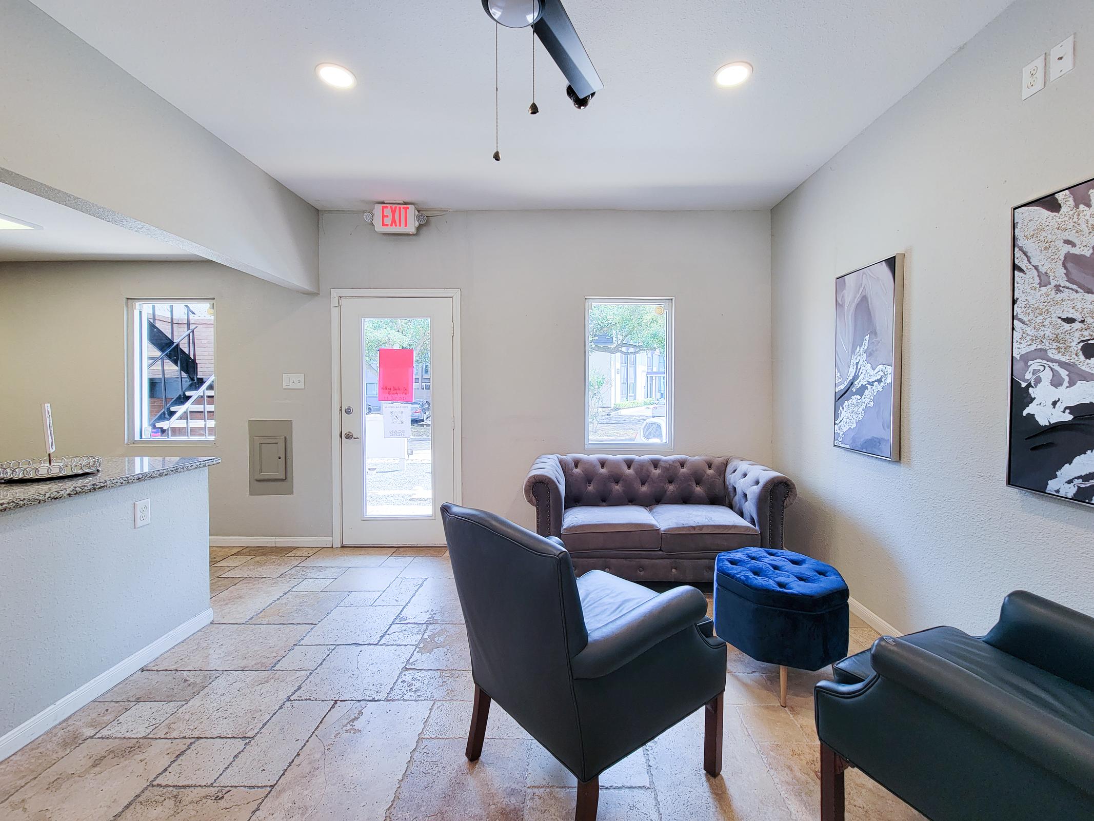 Interior view of a modern lounge area featuring a tufted brown sofa, a blue ottoman, and two stylish black chairs. The walls are painted light gray, and there are two windows providing natural light. The flooring is tiled, and a doorway leads outside.