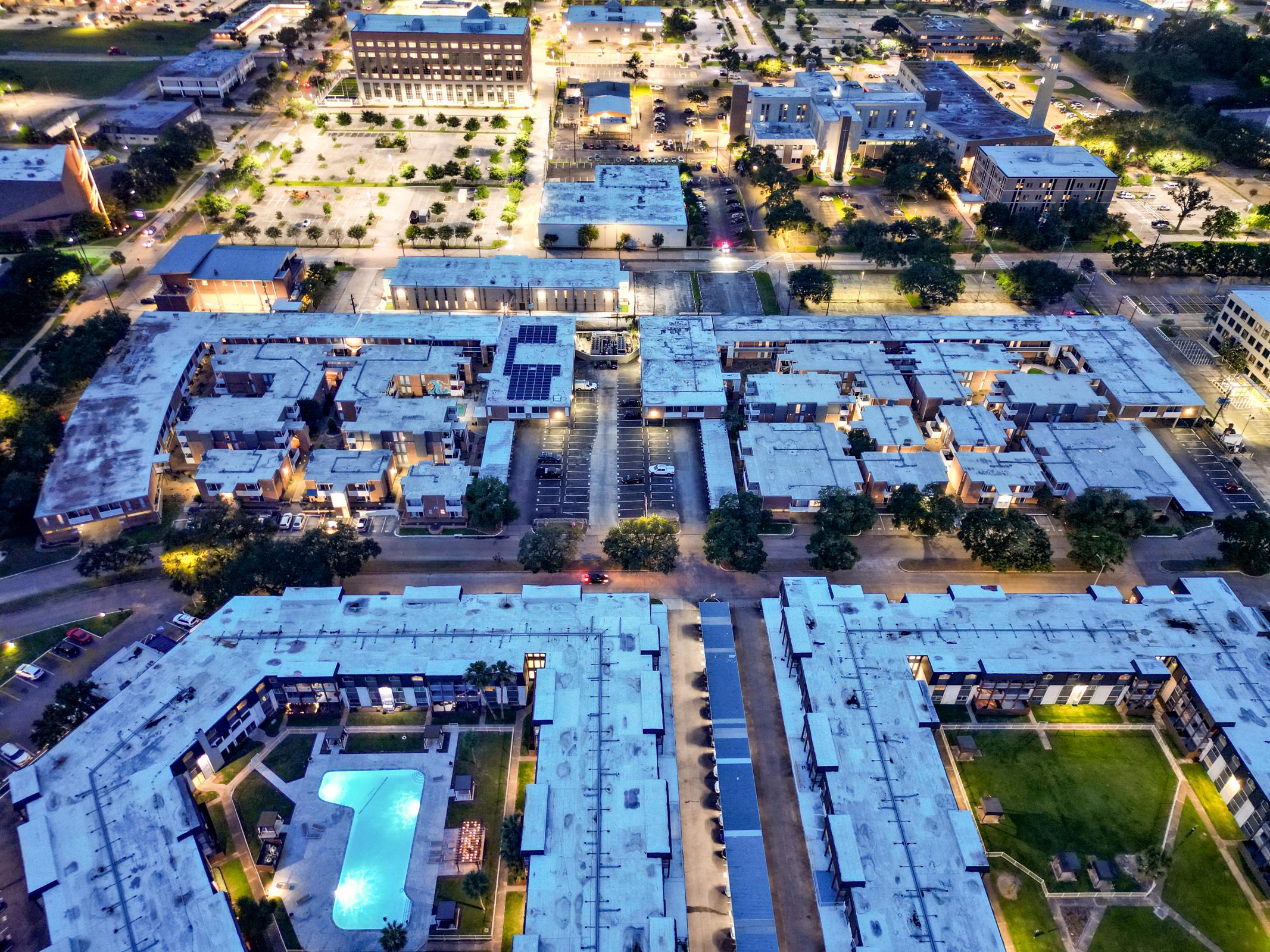 Aerial view of a modern urban residential complex at night, featuring multiple low-rise buildings with flat roofs. The layout includes a central green space and a swimming pool, surrounded by parked cars and illuminated pathways. Nearby, there are additional buildings and a spacious parking area.