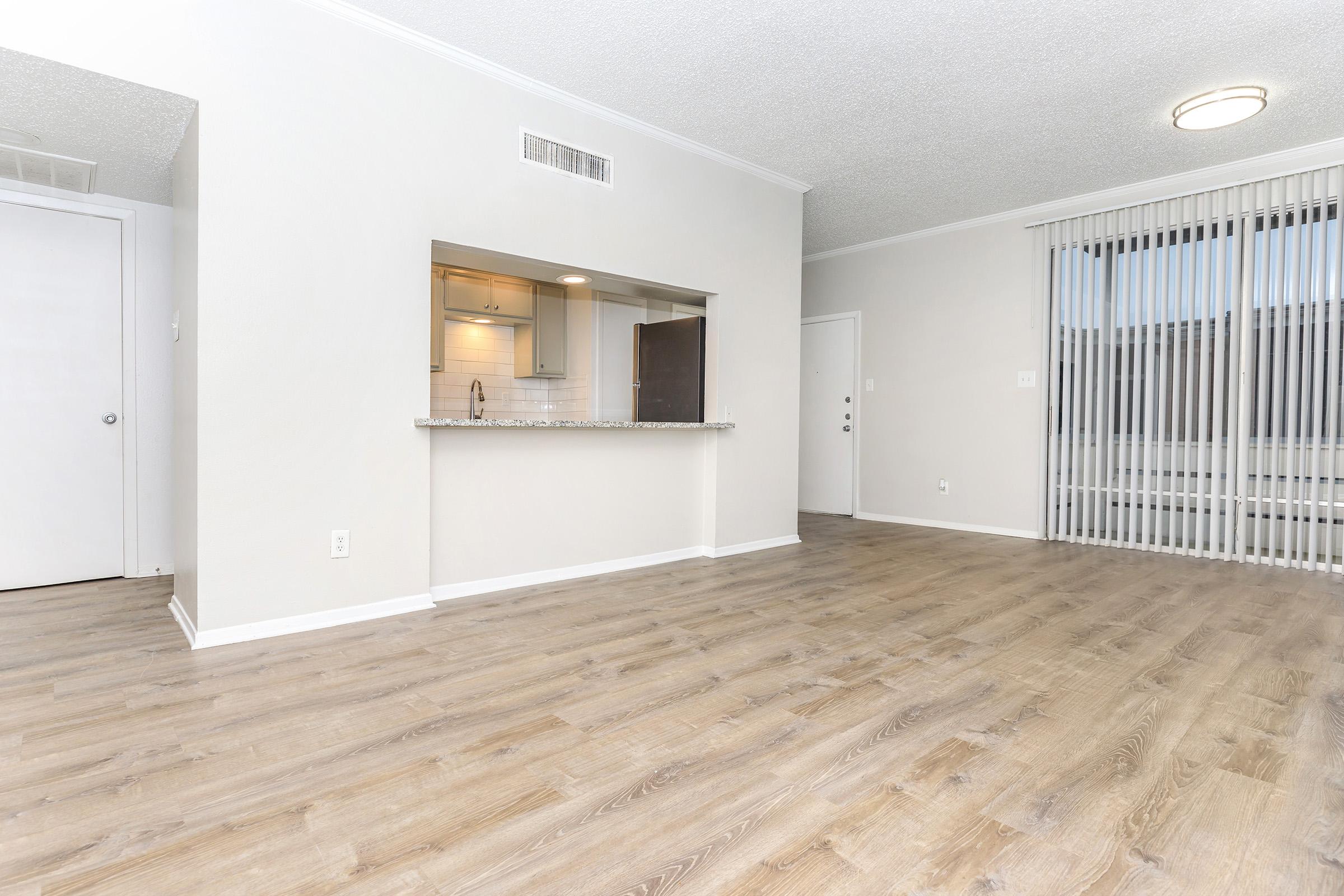 Spacious interior of an apartment featuring hardwood floors, neutral-colored walls, and a modern kitchen area with a granite countertop. Natural light comes in through large windows with vertical blinds. The layout includes an open living space and a door leading to another room.