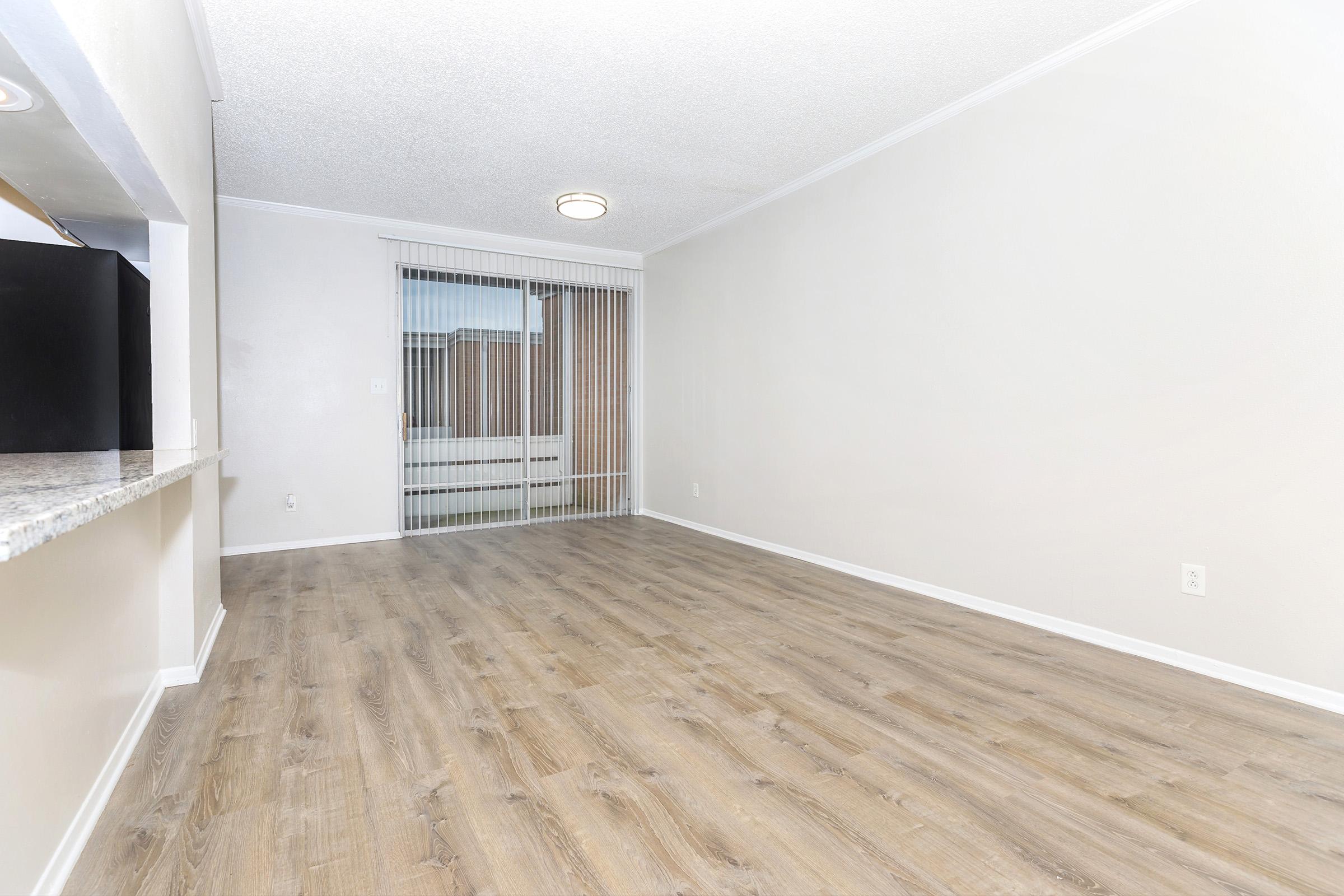 Spacious, empty living room with light-colored walls and wood laminate flooring. A sliding glass door leads to a balcony or outdoor area, allowing natural light to enter. The room features a ceiling light fixture and a small kitchen area visible in the background.