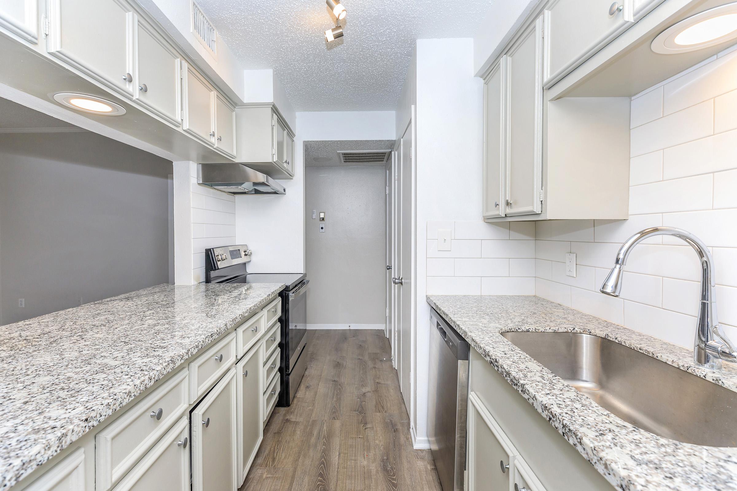 Modern kitchen featuring light gray cabinets, granite countertops, and stainless steel appliances. The space includes a double sink, a stove, and a refrigerator area. The backsplash is white subway tiles, and the flooring is a light wood laminate. Bright lighting overhead enhances the clean, contemporary look.