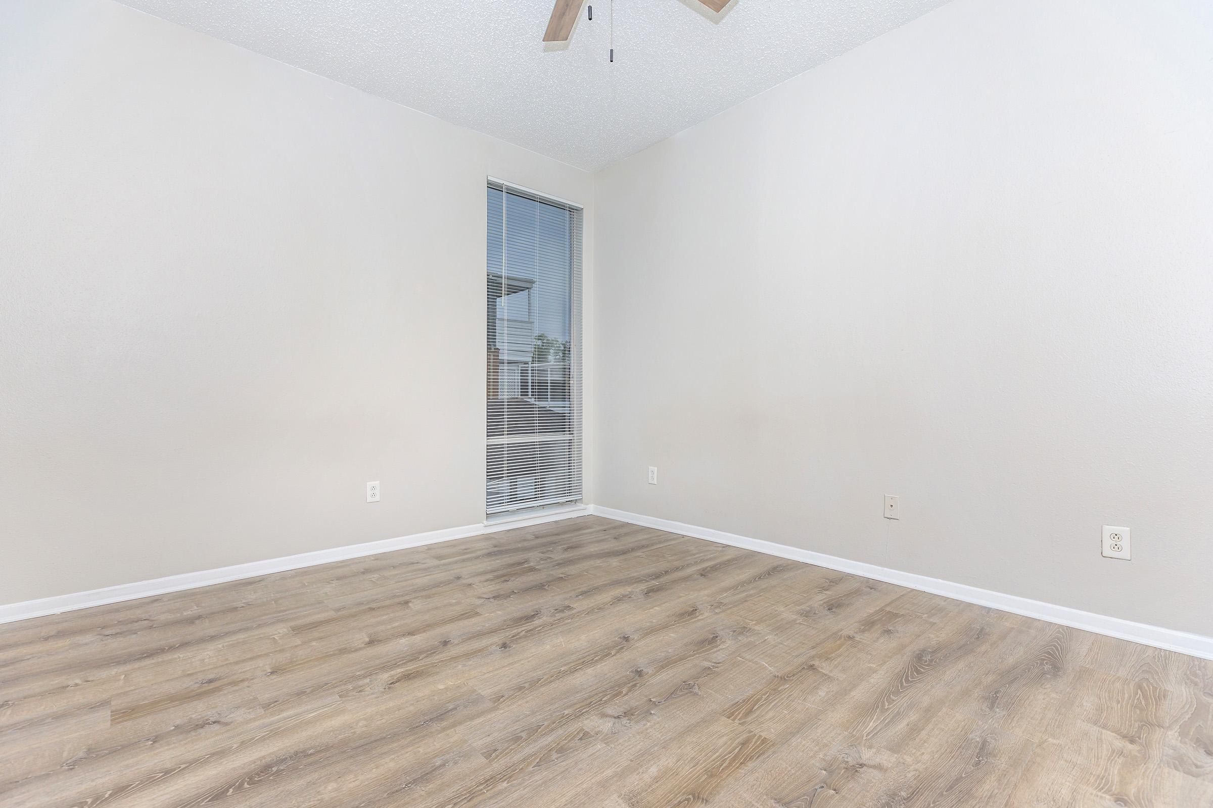 Empty room with light-colored walls and wooden flooring. A ceiling fan is visible, and there is a window with blinds on one wall. The space appears bright and minimalistic, ideal for various uses or decorating.