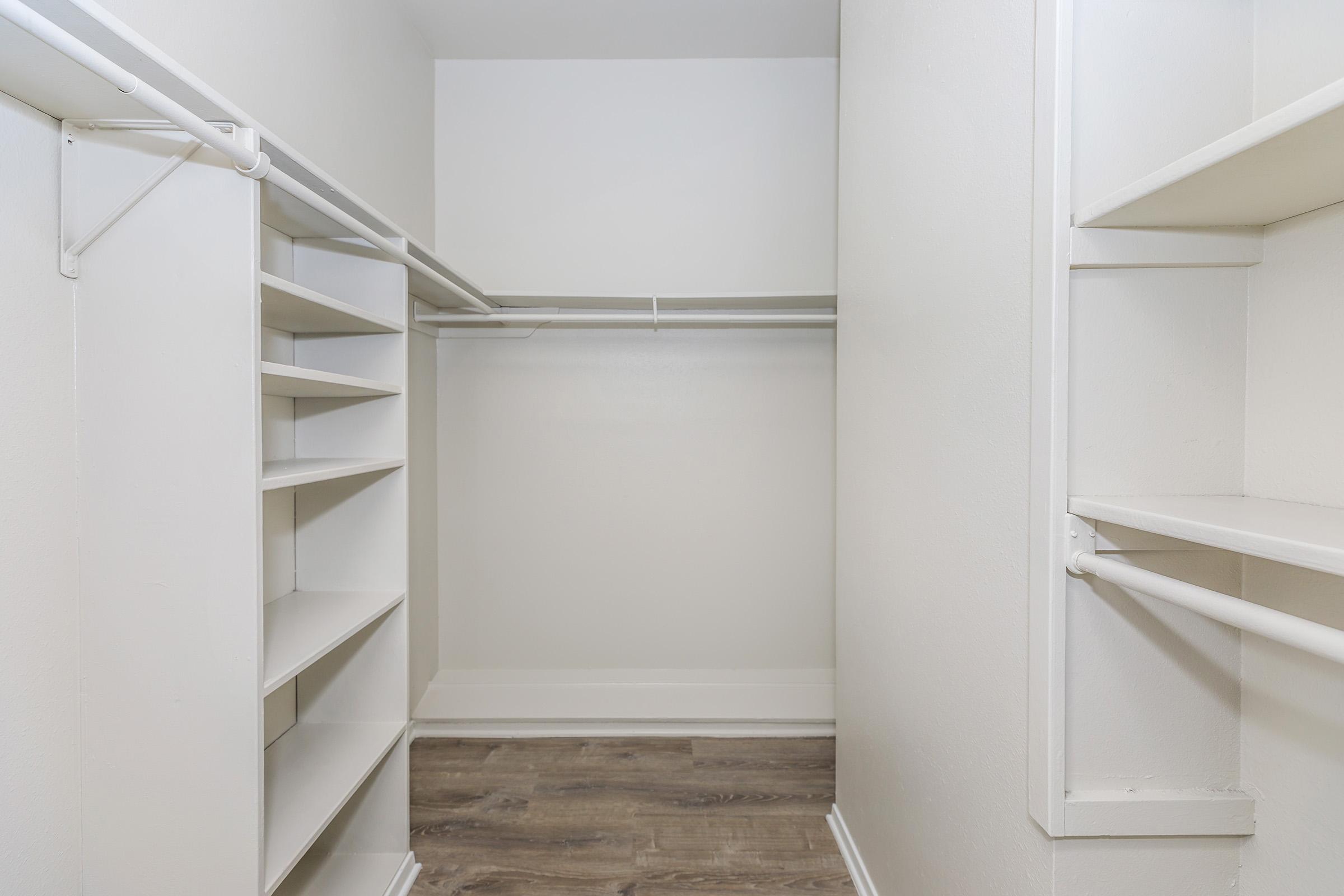 A spacious, empty closet featuring white shelving units along one side, hanging rods for clothes, and light-colored walls. The flooring is a light wood laminate, creating a clean and modern look.