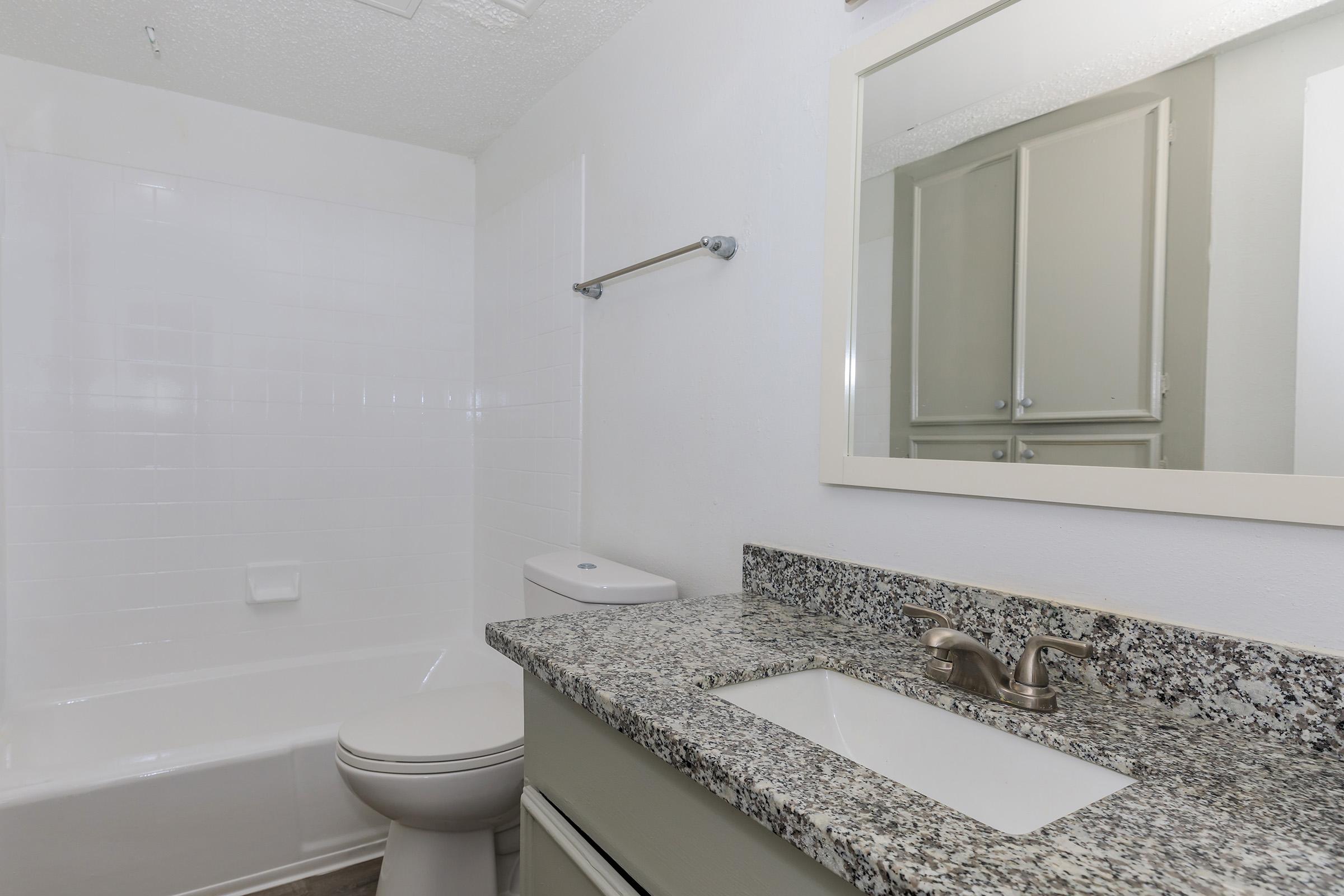 A clean and modern bathroom featuring a white bathtub with a shower, a granite countertop with a sink, a mirror, a toilet, and light-colored cabinetry. The walls are painted white, creating a bright and open space.