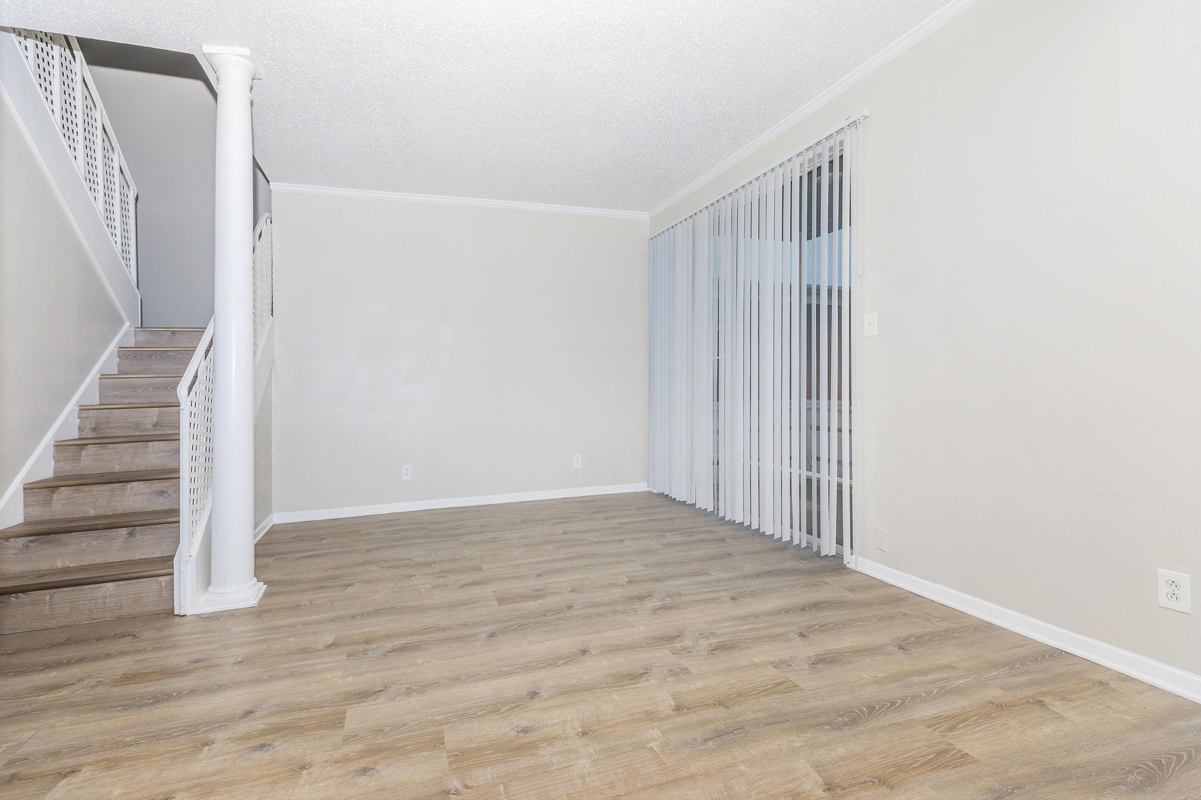 A spacious, empty room with light-colored walls and a wood-like floor. A staircase is visible on the left, and large vertical blinds cover a window at the far end, allowing natural light to enter. The space is well-lit and designed for a welcoming atmosphere.