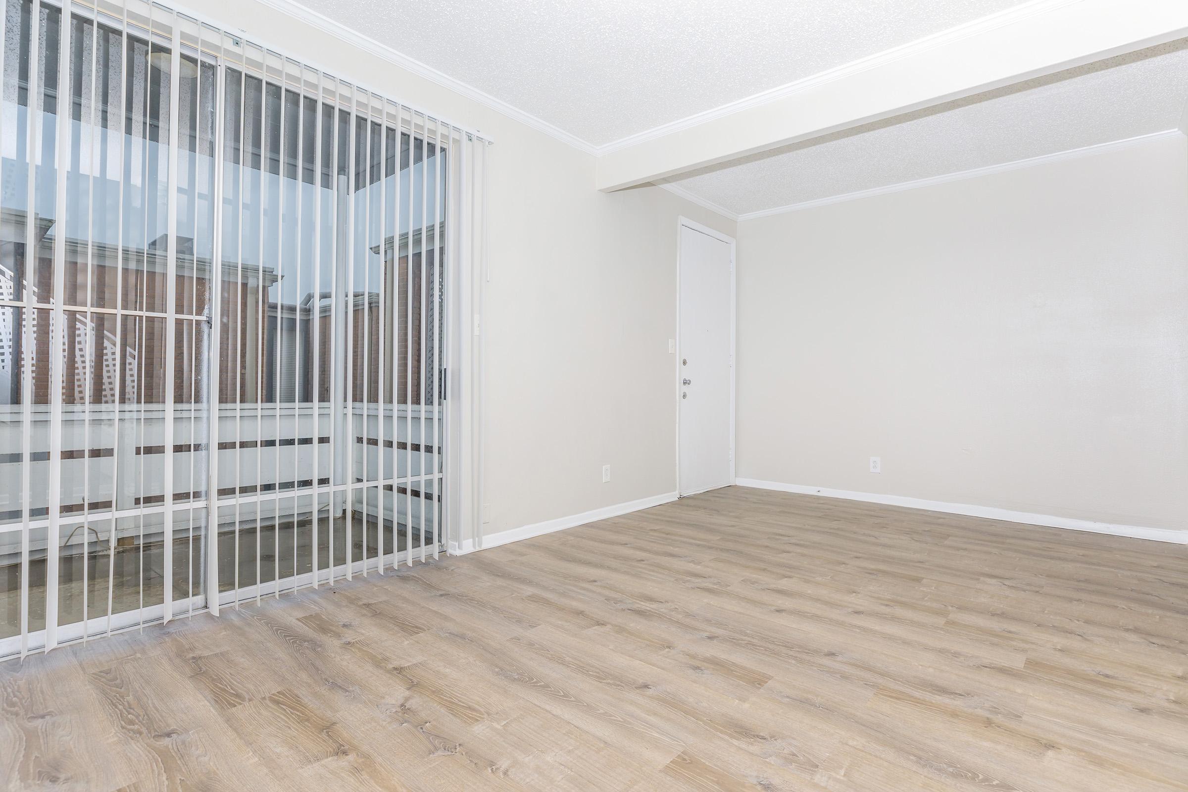 An empty room with light-colored walls and wooden flooring. On one side, there are sliding glass doors with vertical blinds leading to a balcony. The opposite wall features a plain entry door. The space is bright and spacious, perfect for various interior design options.