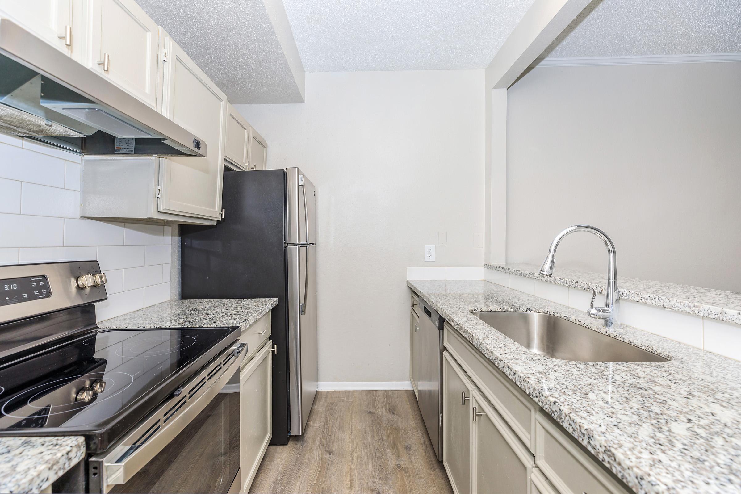 Modern kitchen featuring gray cabinets, stainless steel appliances including a refrigerator and oven, a granite countertop, and a sleek sink. The space has a neutral wall color and a clean, organized layout, ideal for cooking and entertaining. Natural light enhances the inviting atmosphere.
