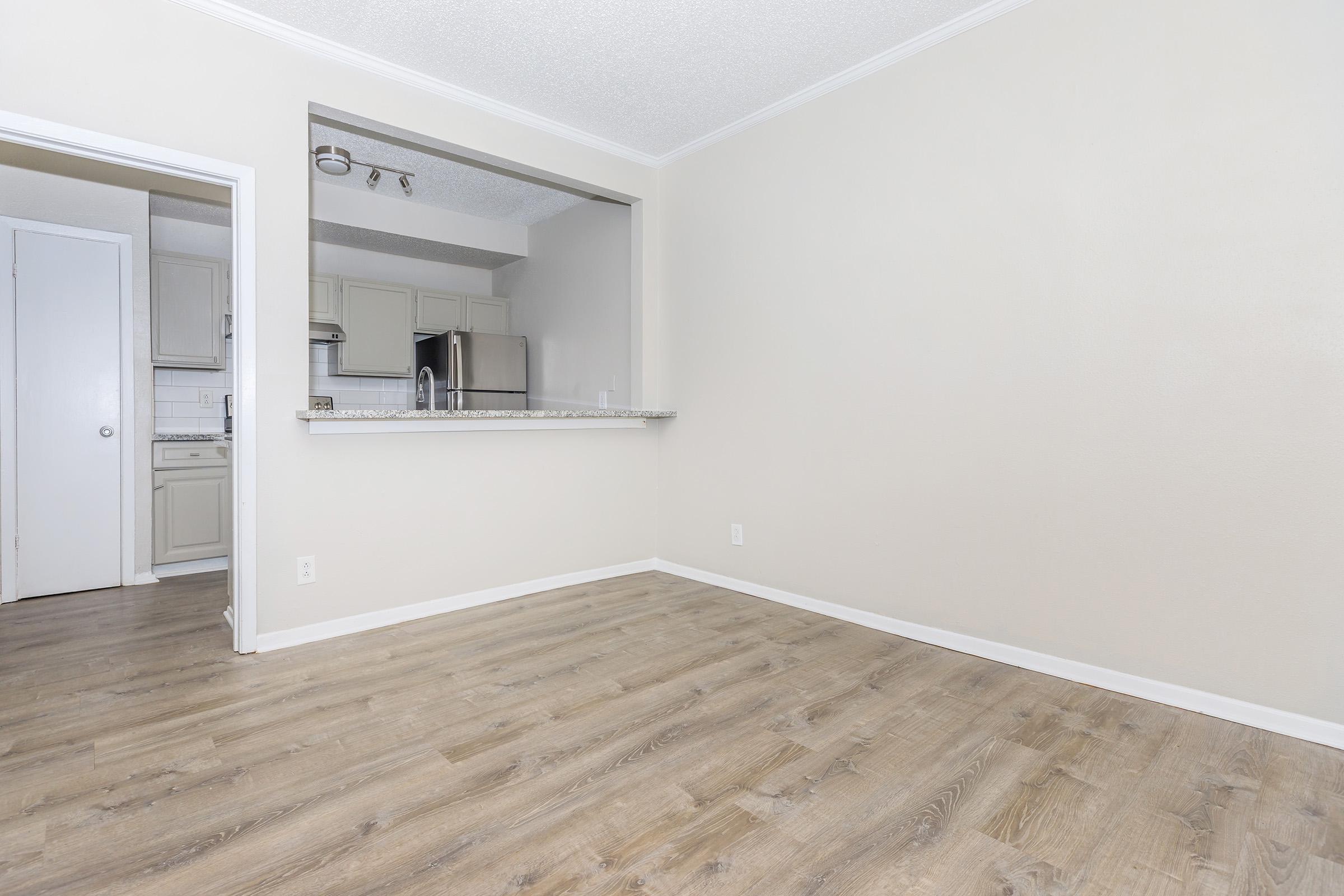 A spacious, empty room with light-colored walls and a wood-like floor. There's an open doorway on the left leading to a kitchen area, visible through a pass-through. The kitchen is equipped with appliances, including a refrigerator and a stainless steel microwave, and is designed with neutral tones.