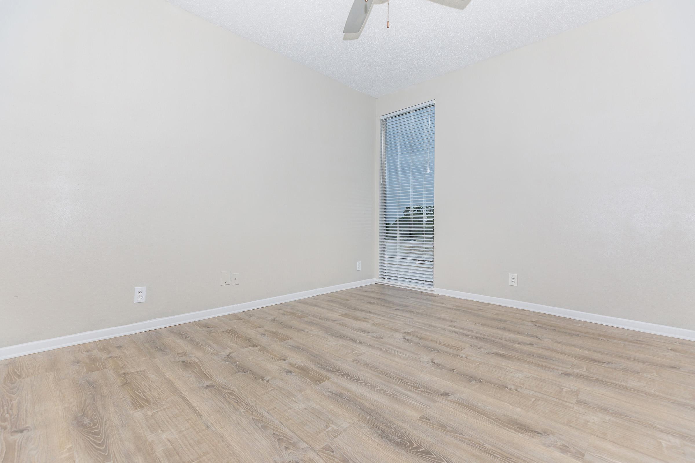 Empty room with light-colored walls and wood-style flooring. A window with blinds lets in natural light, and a ceiling fan is visible. The room is unfurnished, giving it a spacious and minimalist feel.