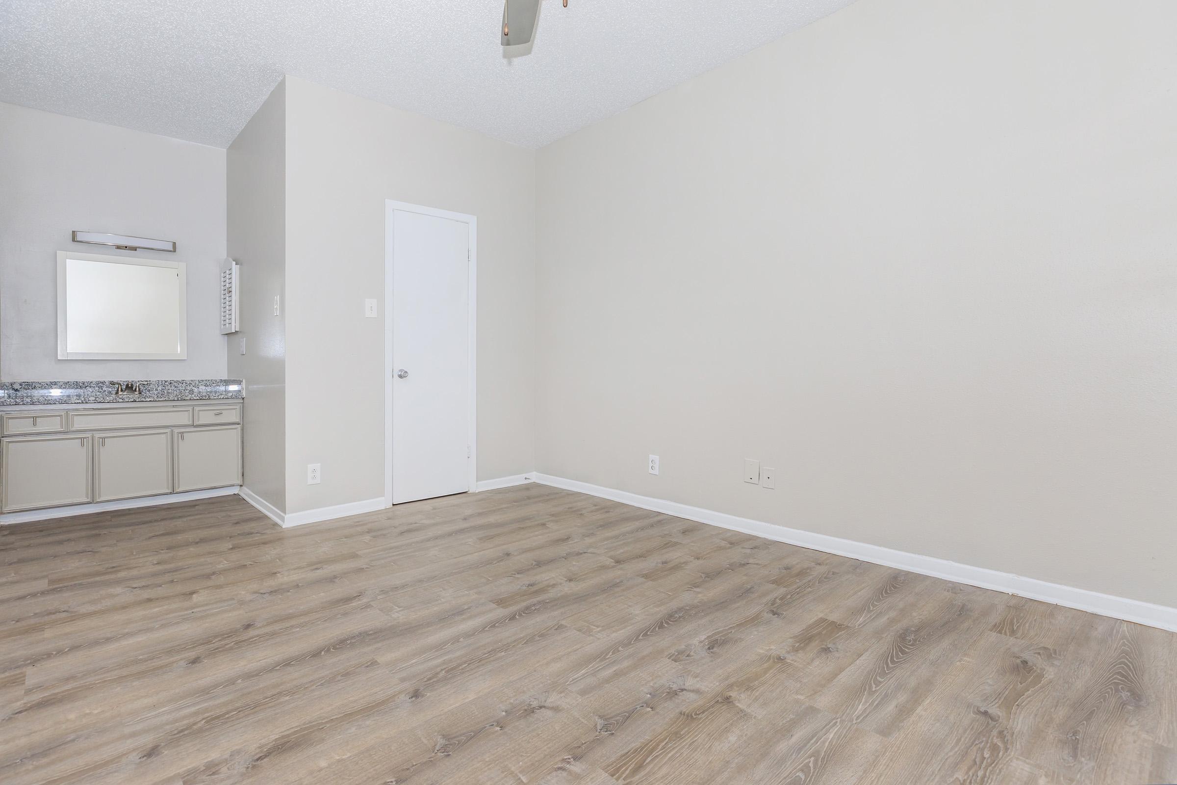 Empty room with light wooden flooring, a small area with a mirror and sink on the left, and a white door on the right. The walls are painted in a neutral color, creating a spacious and bright environment. The lighting is soft, enhancing the room's airy feel.