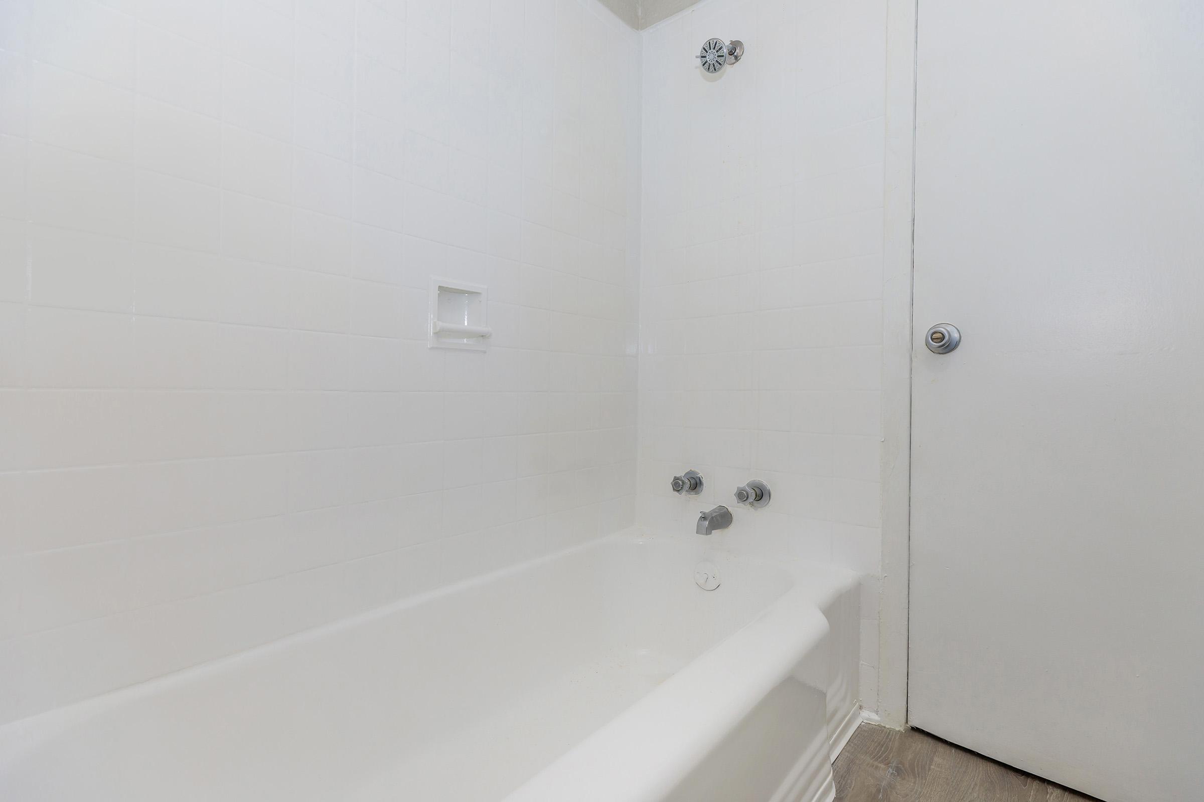 A clean, empty bathroom featuring a white bathtub with chrome faucets, set against white tiled walls. A plain door is visible on the right, adding to the minimalist and bright aesthetic of the space.
