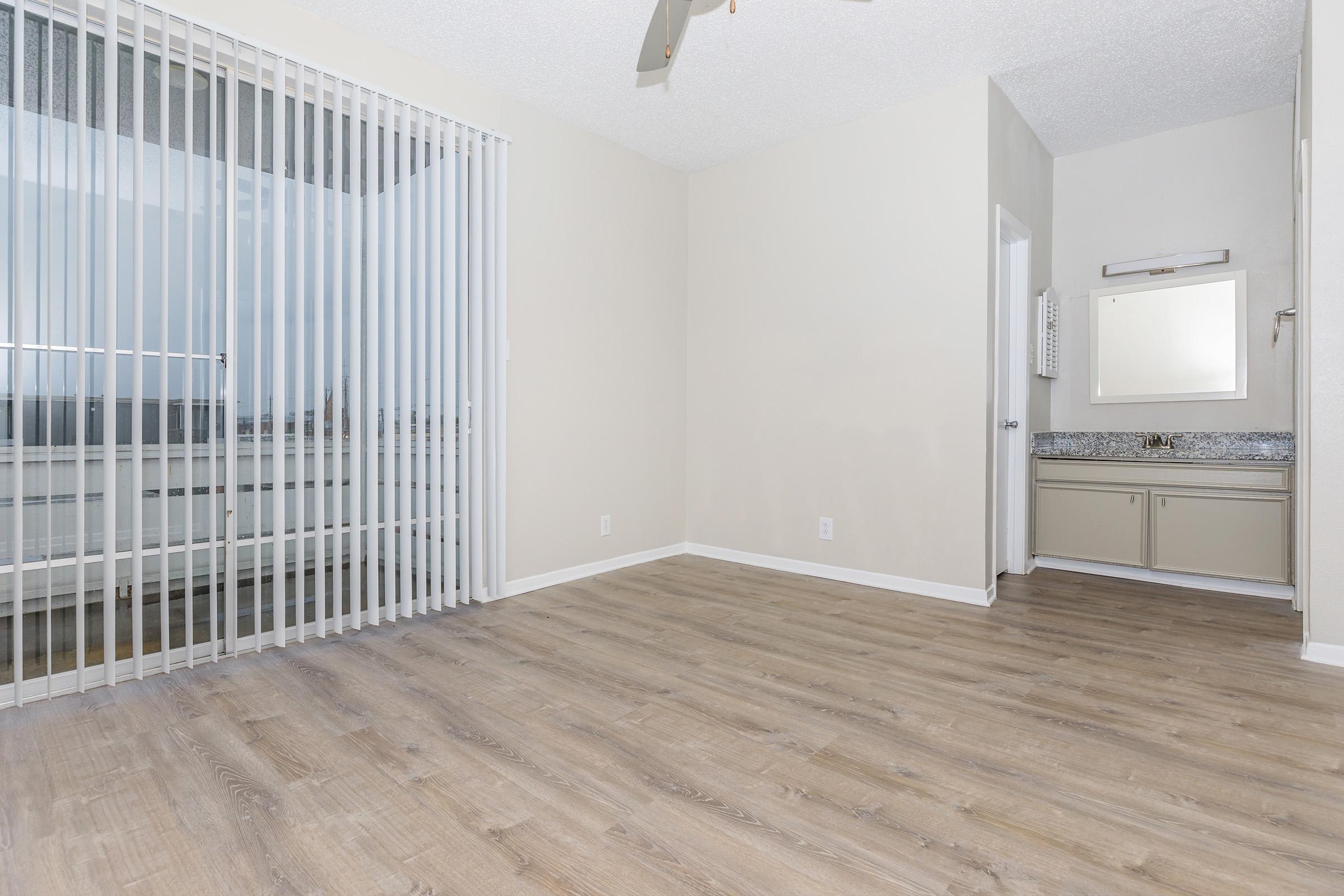 A spacious, empty room with light-colored walls and a wooden floor, featuring a sliding glass door with vertical blinds. On one side, there's a small area with a mirror and countertop. Natural light is coming in from the window, creating a bright and inviting atmosphere.
