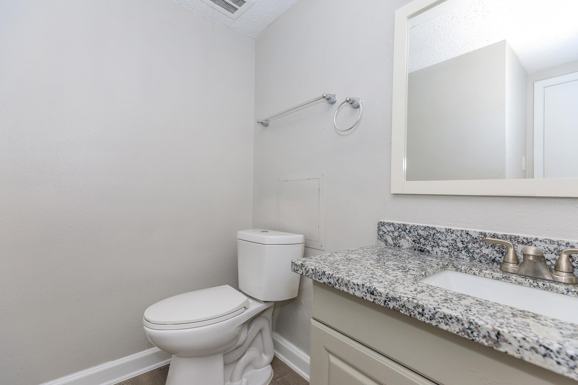 A small, modern bathroom featuring a white toilet, a countertop with a granite surface, a sink with two faucets, and a simple mirror. The walls are painted in a light color, and there's a towel bar on the wall. The floor is tiled, contributing to a clean and minimalistic aesthetic.