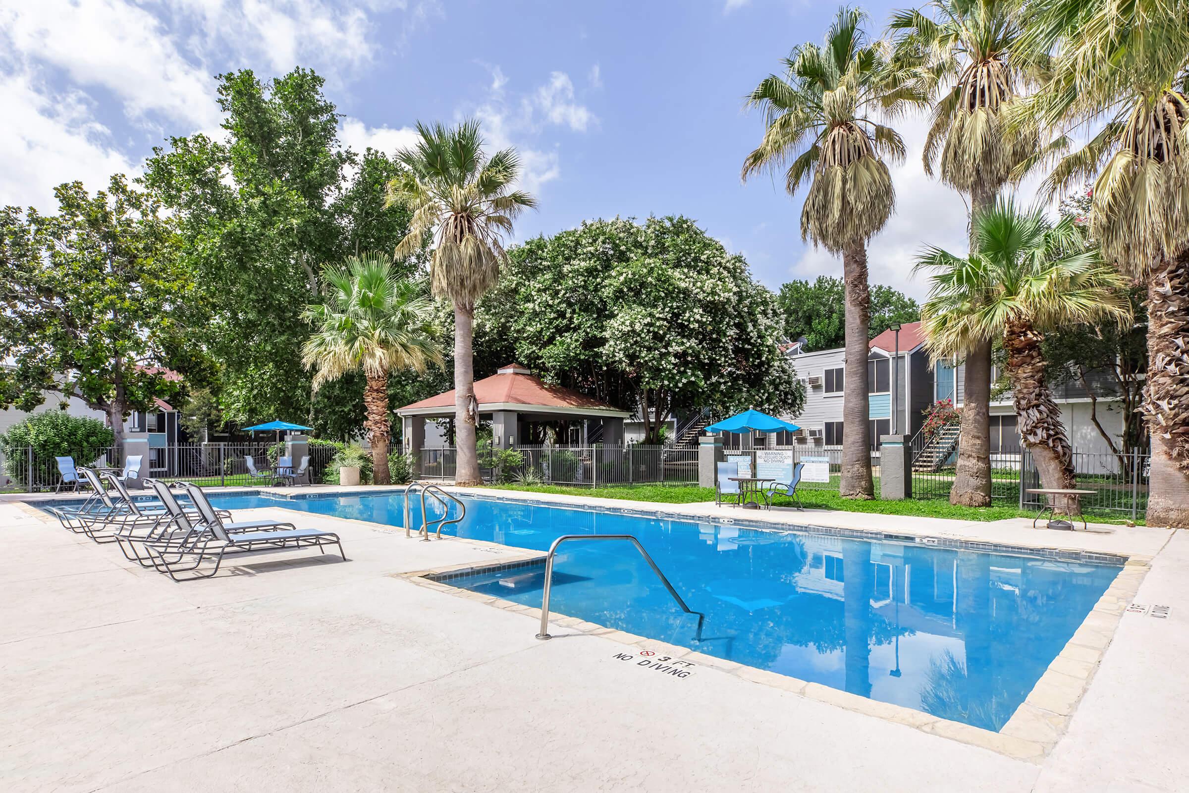 A clear blue swimming pool surrounded by palm trees and lounge chairs, with a gazebo in the background. Blue umbrellas provide shade, and there are several residential buildings visible nearby. The sky is partly cloudy, creating a bright and inviting atmosphere.