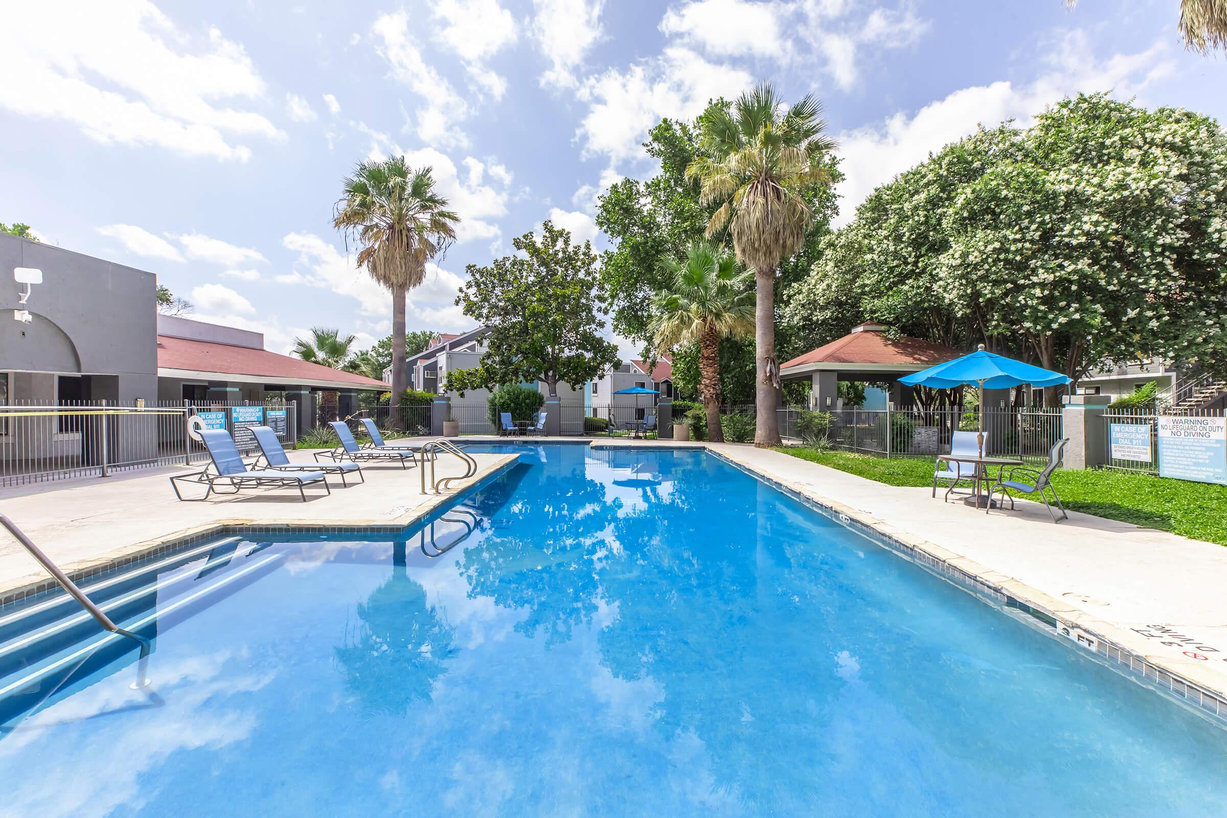 A clear blue swimming pool surrounded by palm trees and lounge chairs. There are shaded seating areas and a few buildings in the background. The sky is partly cloudy, creating a bright and inviting atmosphere.