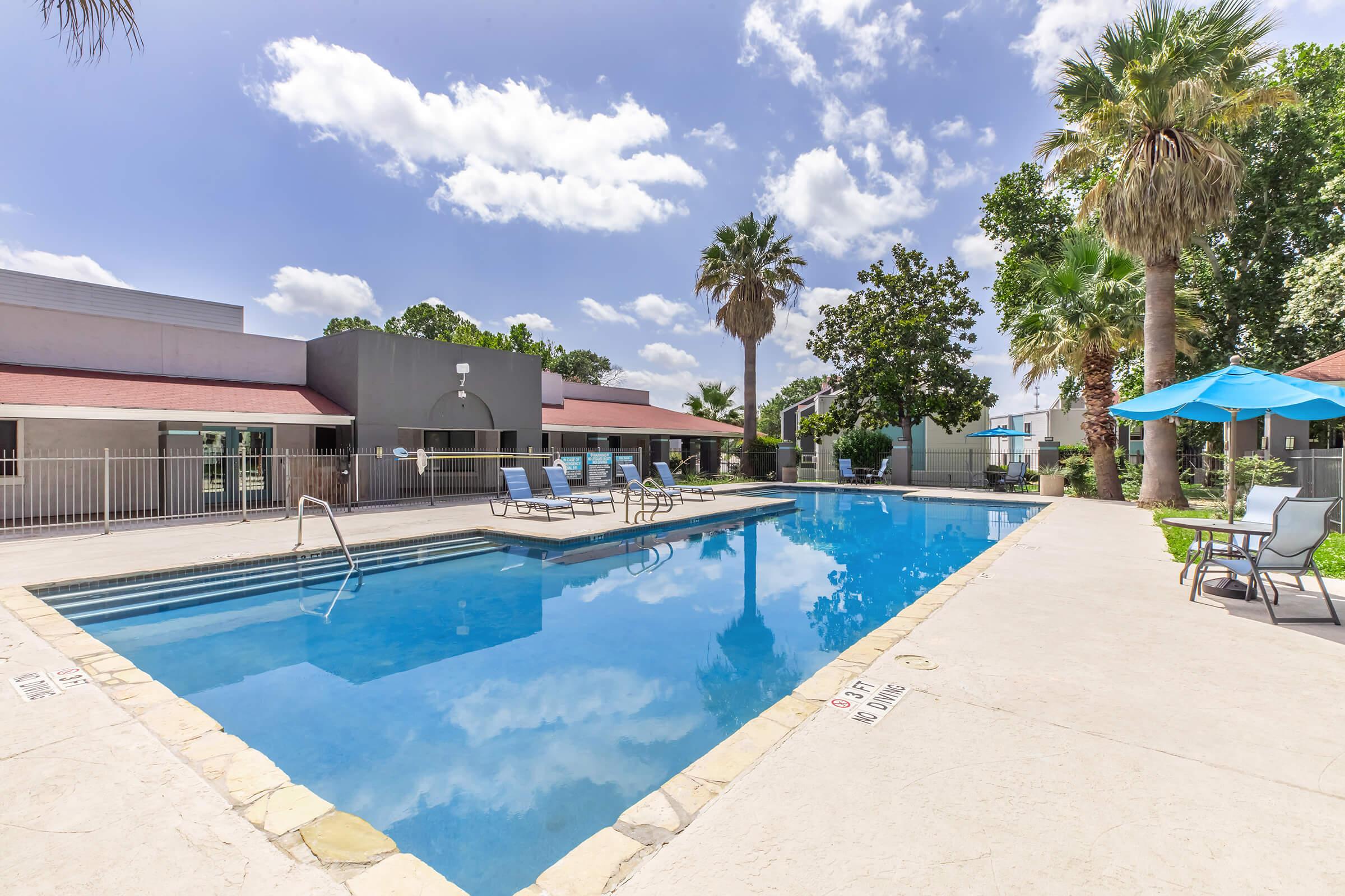 A clear blue swimming pool surrounded by lounge chairs under a bright sky with scattered clouds. Palm trees and greenery are visible in the background, alongside a shaded seating area with a blue umbrella. The pool area is well-maintained and inviting.