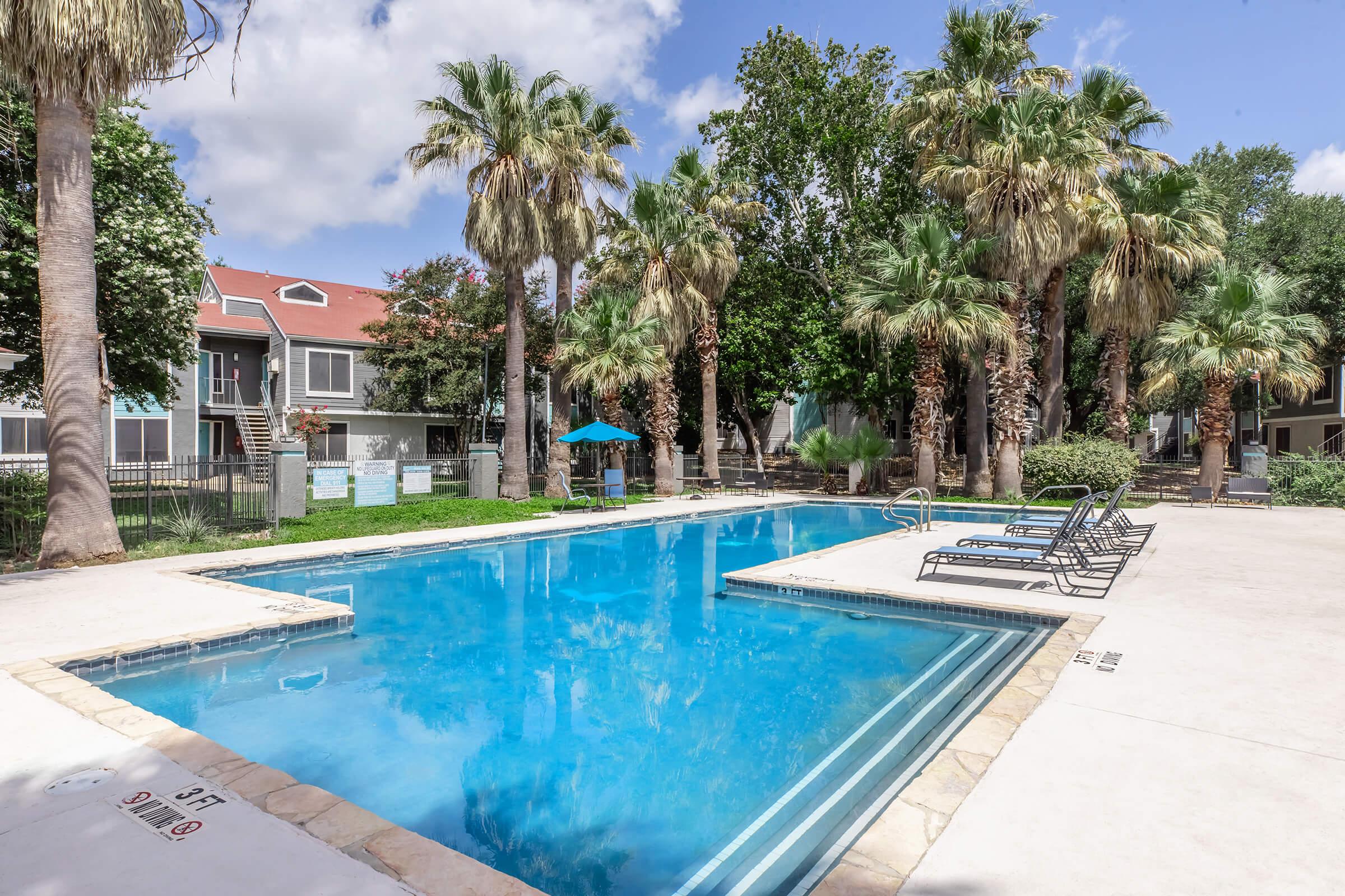 A clear swimming pool surrounded by tall palm trees and lounge chairs. In the background, there are two-story buildings with dark and light exteriors. The sky is bright with a few clouds, creating a vibrant atmosphere in a relaxing outdoor setting.