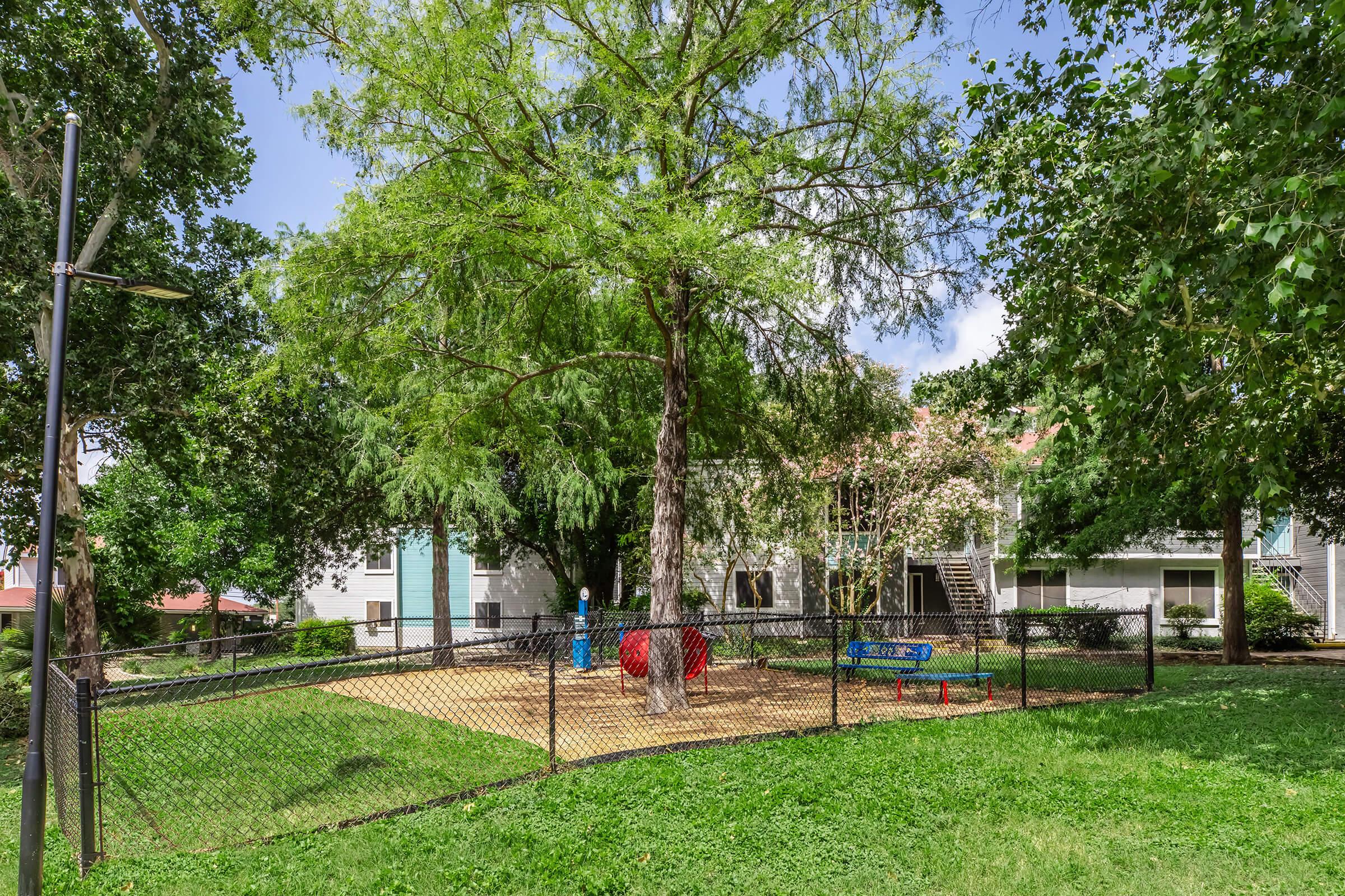 A playground area surrounded by a fence, featuring a red and blue play structure under a large tree. Grass surrounds the play area, and several residential buildings are visible in the background, along with a clear blue sky.