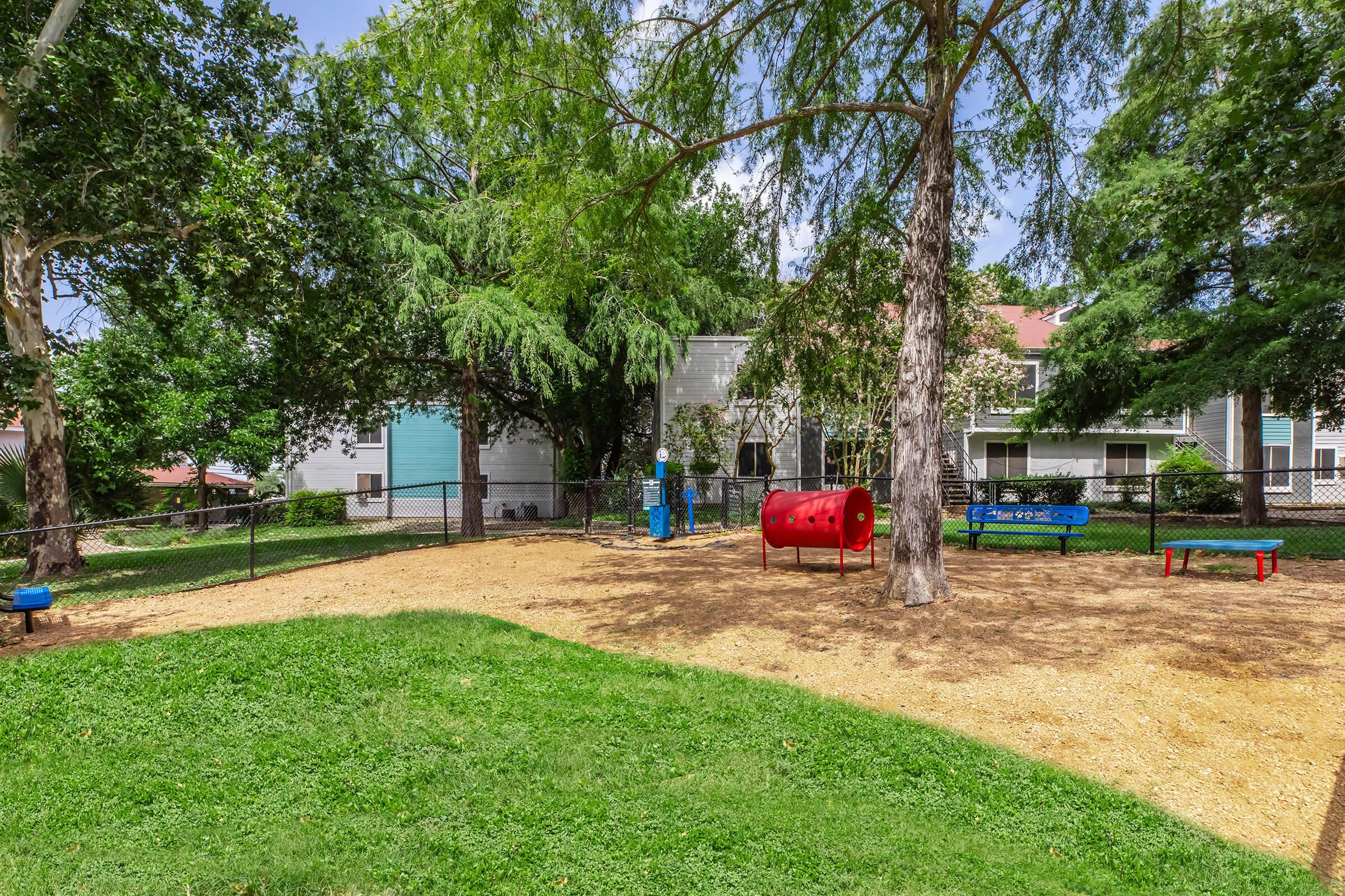 Children's playground area featuring a red tunnel slide and blue benches, surrounded by trees and grass. Residential buildings are in the background, with a fence enclosing the play area. The scene is bright and inviting, perfect for outdoor play.