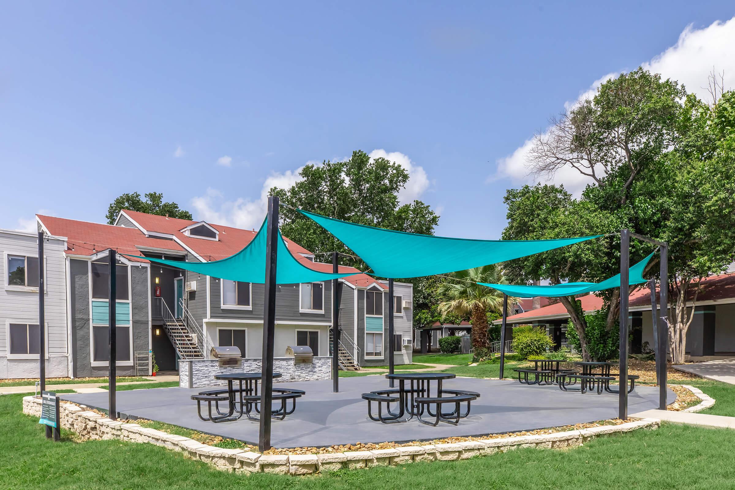 Outdoor area with large shade sails providing cover over several picnic tables. In the background, there are modern apartment buildings surrounded by greenery under a clear blue sky. The scene is bright and inviting, perfect for relaxation or social gatherings.