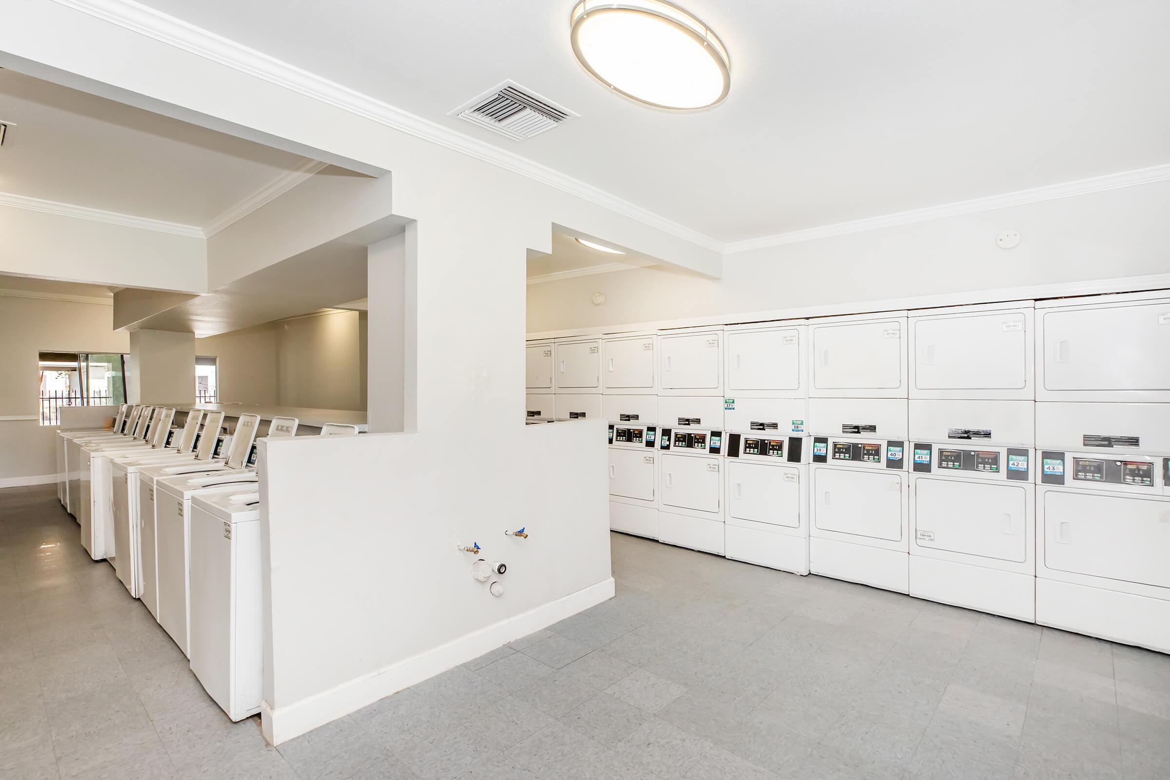 A bright and clean laundry room features rows of white washing machines and dryers against a light-colored wall. The floor is tiled in a light shade, and there is a ceiling light fixture illuminating the space. The atmosphere is modern and well-maintained.