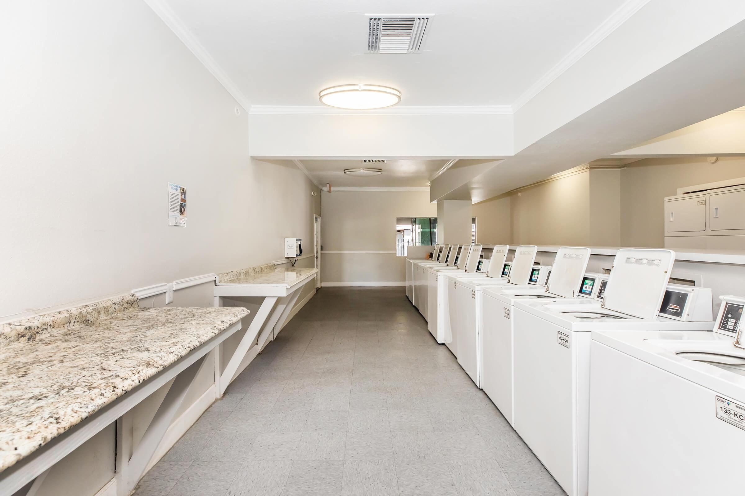 A clean, well-lit laundry room featuring several rows of white washing machines and dryers. There is a granite countertop running along one side, and the walls are painted in neutral tones, creating a spacious and organized environment for doing laundry.