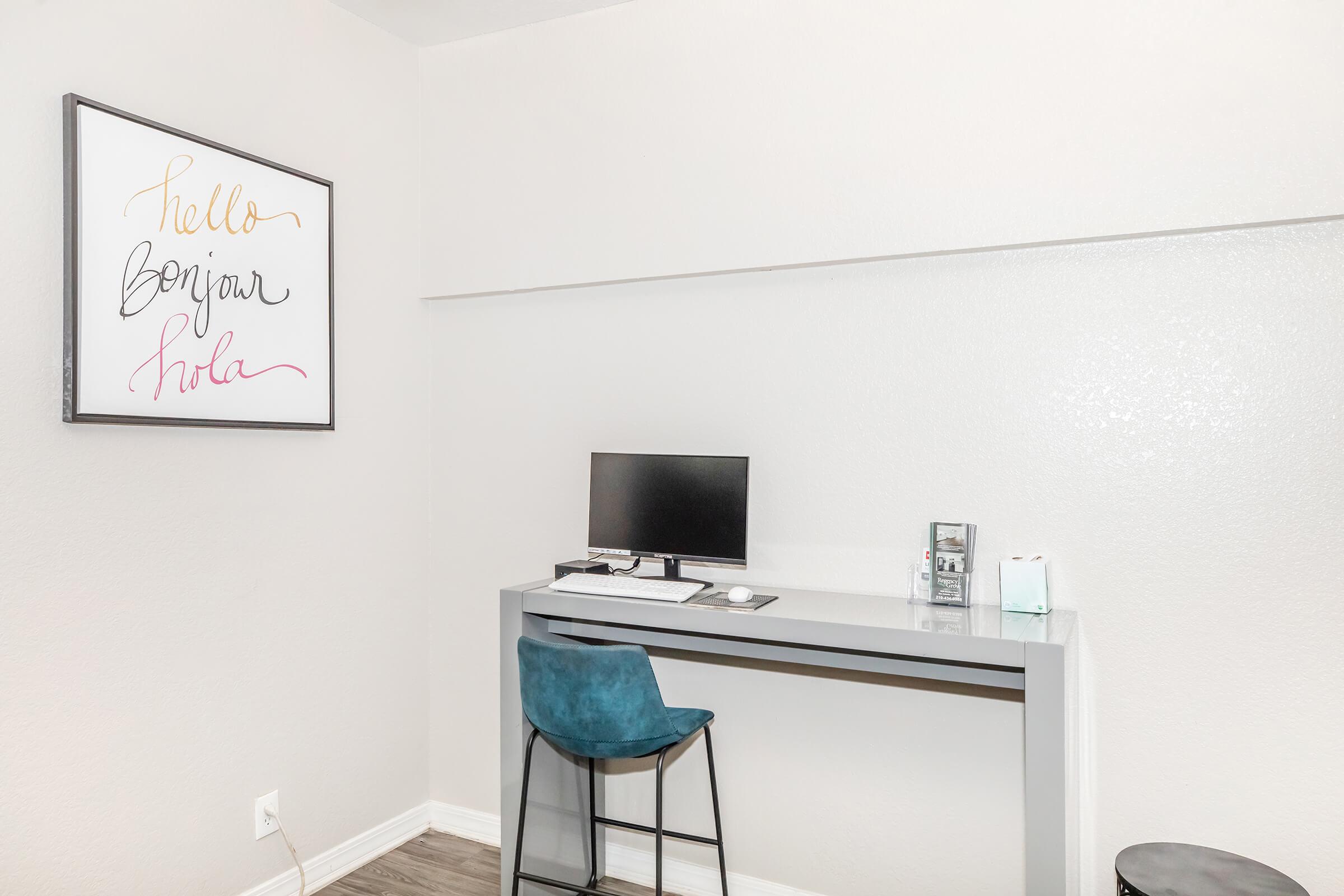 A minimalist workspace featuring a sleek grey desk with a computer monitor, a blue chair, and a framed decorative wall art that says "Hello" in various colors. The wall is plain and light-colored, creating a clean and modern aesthetic.