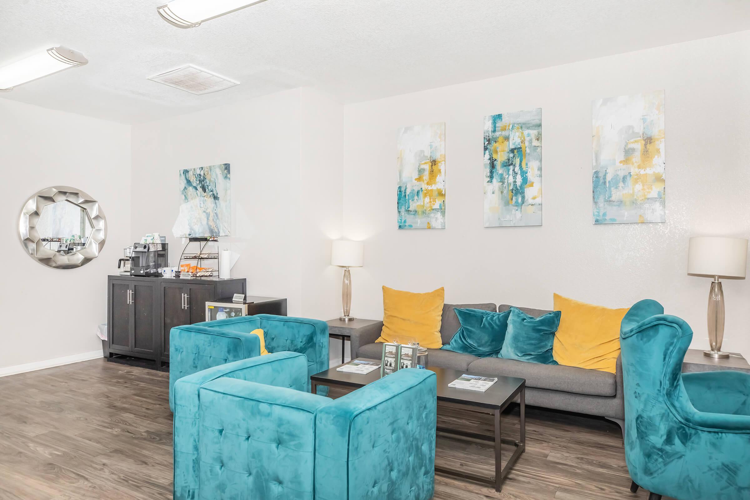 A cozy lounge area featuring turquoise chairs and a gray sofa with yellow cushions. The walls display abstract paintings in blue and yellow tones. A round mirror is on one wall, and a side table with a coffee station is visible, creating a welcoming and modern atmosphere.